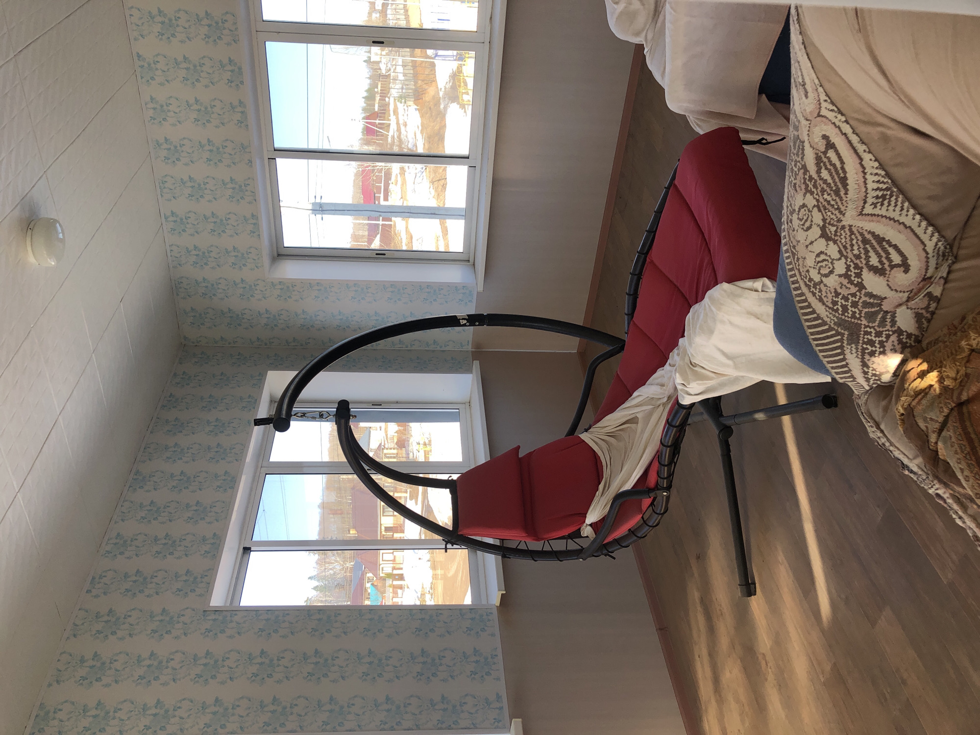 The image shows an interior view of a room in Noviy, Russia. The room has a window with a view of buildings, likely residential, and trees. The walls are decorated with light blue floral patterned wallpaper. The floor is a light wood laminate. A black, curved frame chair with red cushions is positioned near the window. A white blanket is draped over the chair. Part of a sofa with patterned pillows is visible to the right of the chair. There are no people visible in the image.