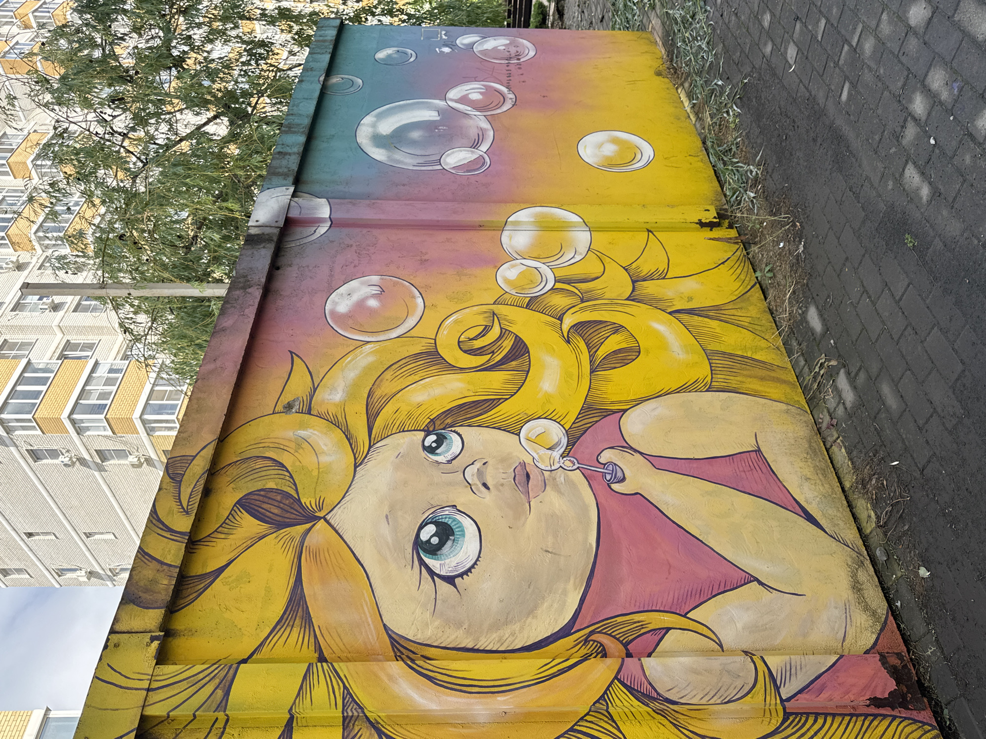 A close-up, tilted shot reveals a vibrant mural adorning what appears to be a utility box or similar infrastructure in Krasnodar, Russia. The artwork depicts a young girl with flowing yellow hair and large, expressive eyes, blowing bubbles. The background transitions from a blue-green hue at the top to a yellow-pink gradient, interspersed with floating bubbles. The mural is positioned next to a paved walkway. A residential building is visible in the background to the left. A tree with green foliage is positioned above the mural.