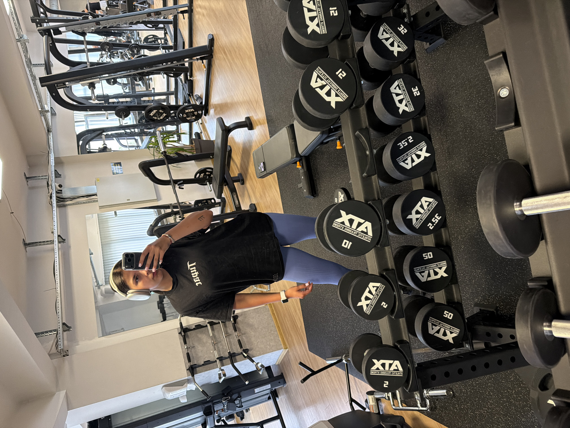 A woman with dark hair and headphones is taking a mirror selfie in a gym. She is wearing a black t-shirt and blue leggings. In the background, there are weightlifting machines, benches, and racks of dumbbells. The gym appears to be modern and well-equipped. The image is taken from a slightly low angle, looking up at the woman.