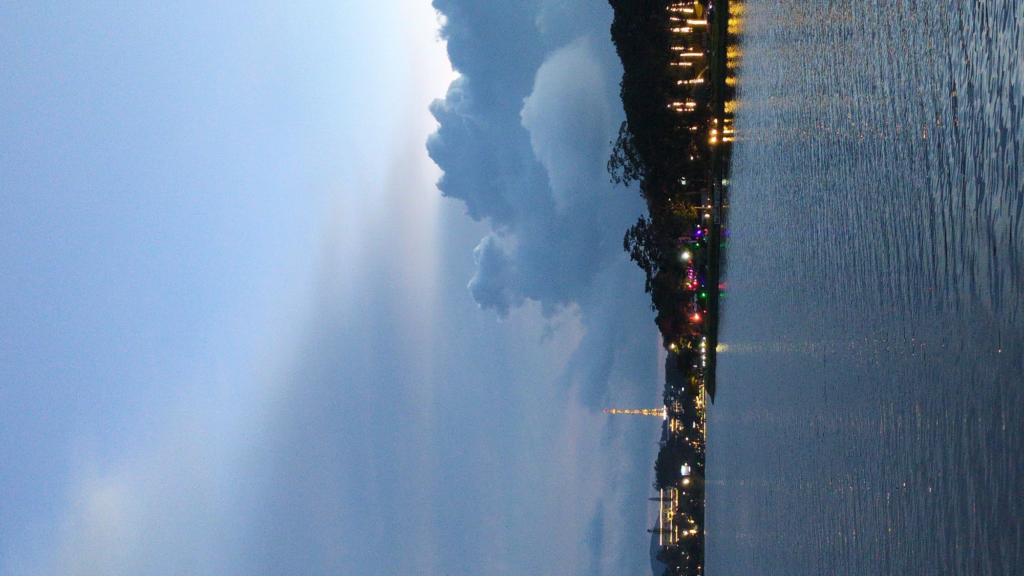 The image depicts Xuan Huong Lake in Da Lat, Vietnam, at dusk. The sky is a gradient of blue and gray, with large, textured clouds dominating the upper portion. The foreground is the calm surface of the lake, reflecting the faint lights from the city and the sky. Along the far shore, a line of trees forms a dark silhouette against the fading light. Several buildings are visible, including a tall, illuminated tower that appears to be a communication or observation tower. String lights and various colored lights are scattered along the waterfront, illuminating trees and possibly market stalls or decorative displays. The overall atmosphere is serene and picturesque, characteristic of a lakeside scene at twilight.