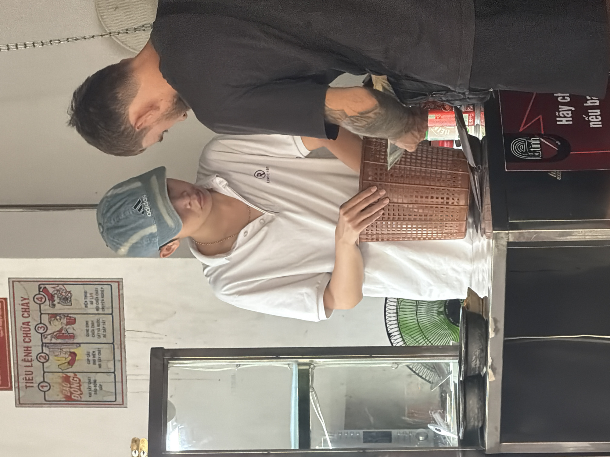 The image captures a close-up view of two individuals in a retail or service setting, likely a food stall or small shop, in Nha Trang Ward, Vietnam. The foreground features a stainless steel counter with two dark bowls and a small dish. Behind the counter, a young person wearing a white polo shirt with a logo and a blue baseball cap is interacting with an adult, who is wearing a black t-shirt and has tattoos on their arm. The adult is holding a basket and what appears to be money or a transaction is taking place. A green electric fan is visible in the background, partially obscured. To the left, a glass-fronted display case contains shelving. A fire safety notice is mounted on the wall above the counter, featuring diagrams and Vietnamese text. The overall atmosphere suggests a casual, everyday transaction.