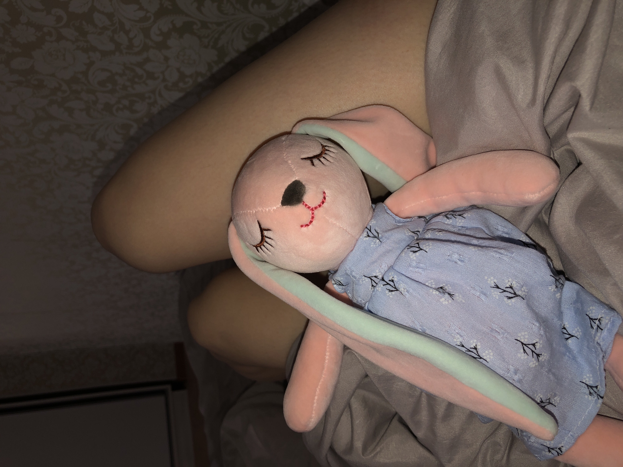 A close-up image shows a pink plush rabbit toy lying on a bed with a light gray bedsheet. The rabbit has a light blue dress with a white floral pattern. The rabbit also has black eyes, a red stitched mouth, and a black nose. The rabbit is close to a person's bare leg, which is positioned near a floral patterned wall. The location of the image is inside a room and there is no location information pertaining to Noviy, Russia.