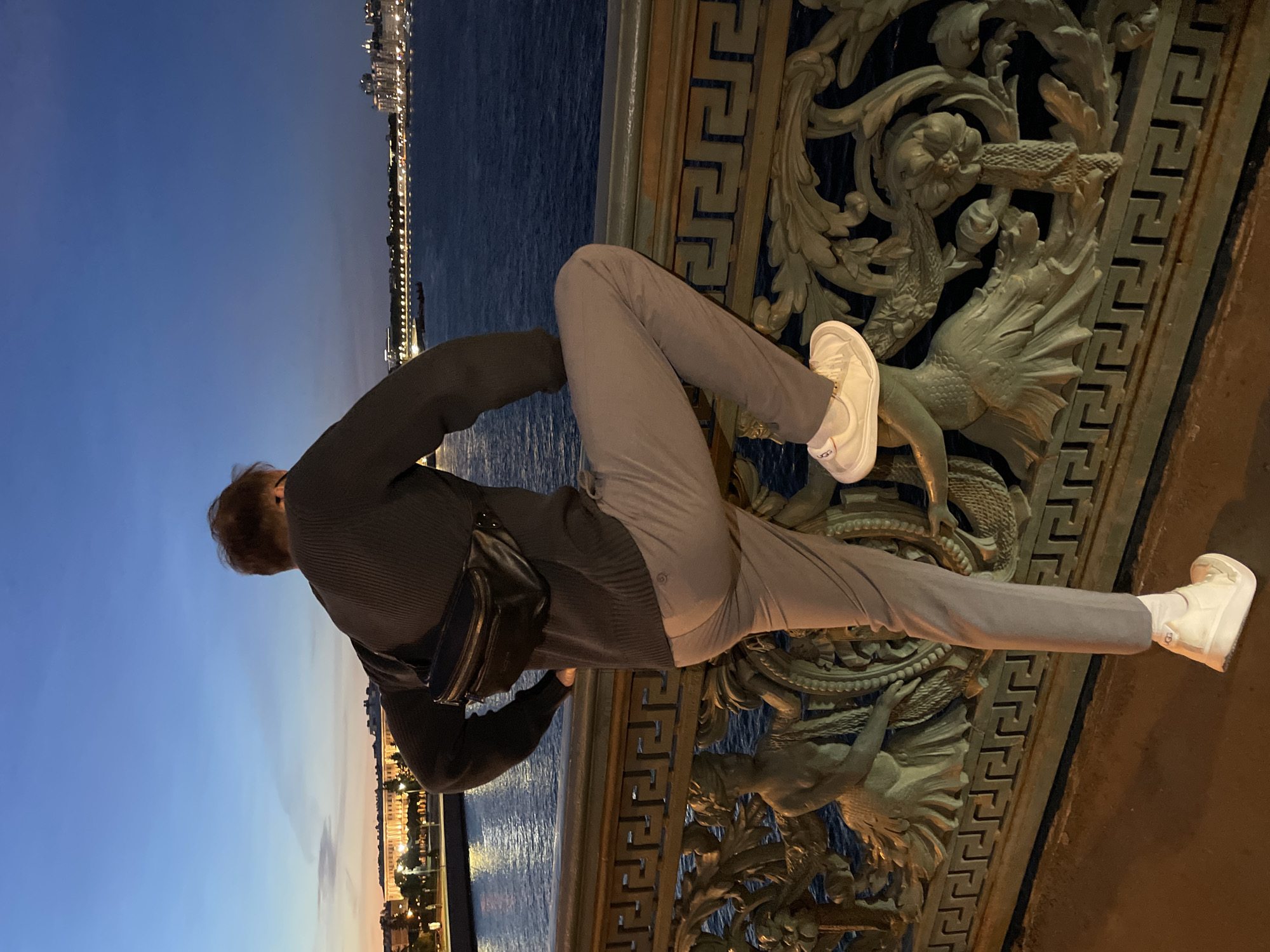 A person is standing on a bridge at dusk, looking out over a body of water towards a city skyline. The person is wearing a dark sweater, light gray pants, and white sneakers. A black fanny pack is worn across the chest. The bridge has ornate metal railings with decorative figures. The sky is a deep blue with hints of orange and pink near the horizon. Lights are visible along the city's waterfront and in buildings. The water is dark and reflects the lights.