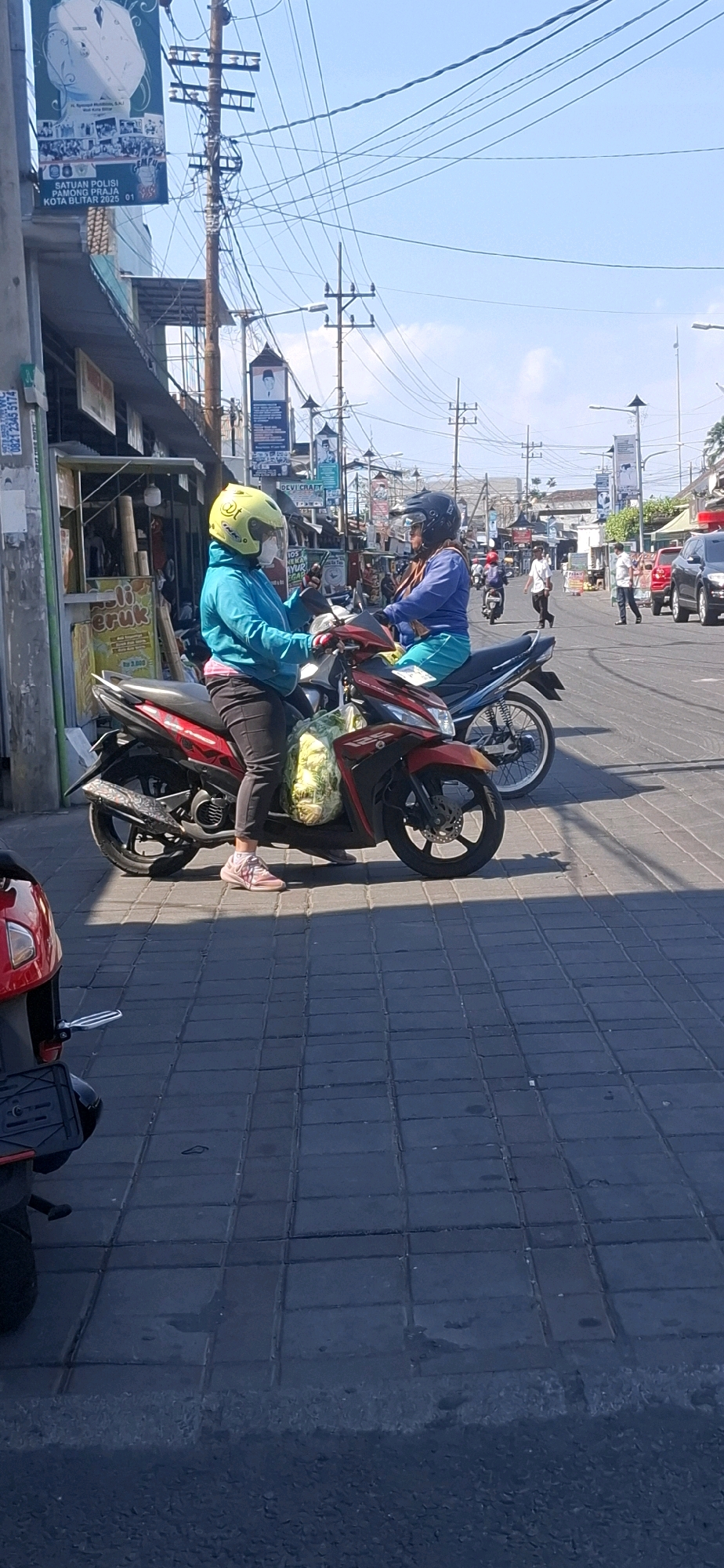 Two individuals are seen on motorcycles in the city of Blitar, Indonesia. The person in the foreground, riding a red motorcycle, is wearing a teal jacket, black pants, pink shoes, a yellow helmet, and a white face mask. They are holding a smartphone in their right hand. A large bag of produce is strapped to the front of the motorcycle. The person behind them, on a blue motorcycle, is wearing a purple jacket and a dark helmet. They are looking forward. The scene appears to be on a paved street with buildings lining the left side and a clear sky above. Utility poles and wires are visible throughout the image. There are other vehicles and pedestrians in the background, suggesting a busy urban environment.