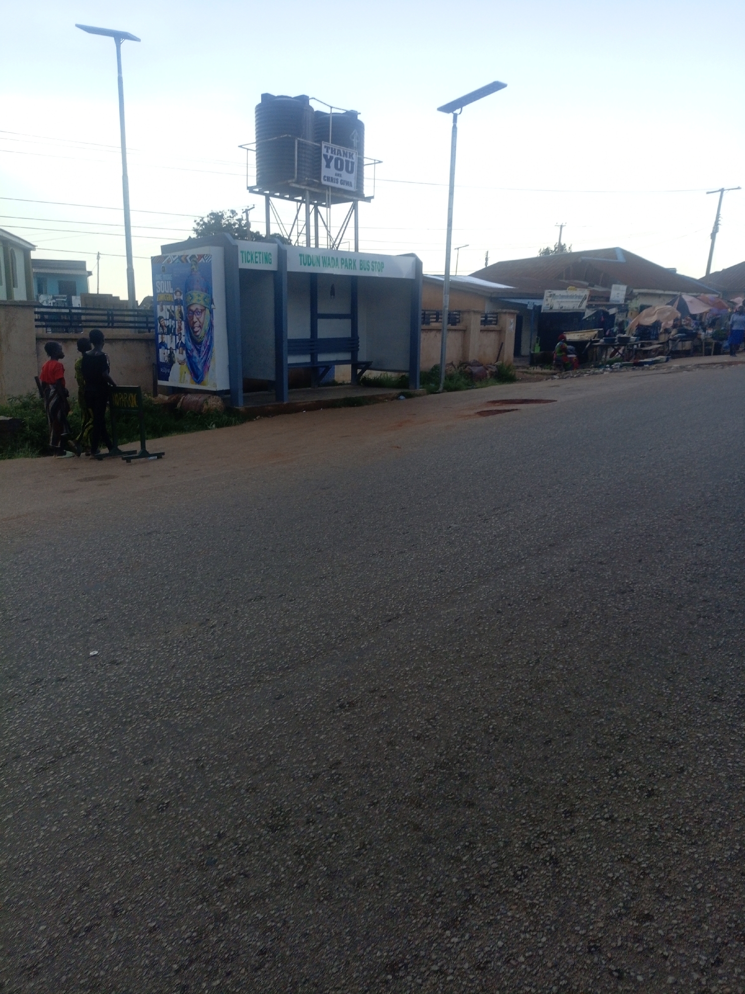 The image shows a street scene in Jos, Nigeria. On the left, a bus stop labeled "TUDUN WADA PARK BUS STOP" is visible. A large poster featuring a portrait of a woman in traditional attire is affixed to the side of the bus stop. To the left of the bus stop, three children are standing on a grassy verge. A solar-powered street light stands tall in the background. The street itself is paved with asphalt and appears to have some water damage or potholes. In the background, several buildings and market stalls with awnings are visible. The sky is clear and bright.