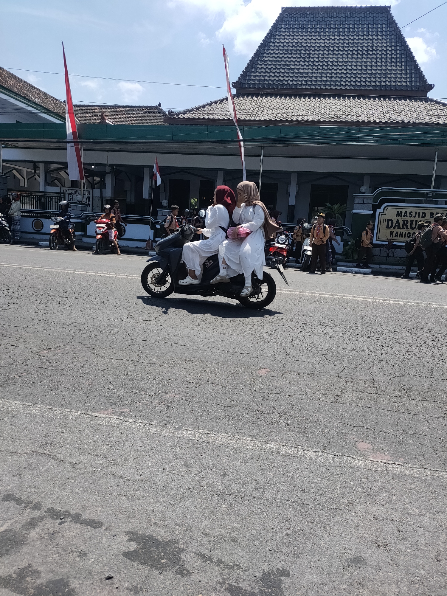 In the city of Kanigoro, Indonesia, on a bright, sunny day, a motorcycle carrying two young women is seen traveling on a cracked asphalt road. The woman in the back, wearing a white hijab and a white dress, is holding a pink bag. The woman in front, also wearing a hijab and a white dress, appears to be driving the motorcycle. In the background, a building with a dark, pitched roof stands prominently. A sign with text "MASJID BESAR 'DARUL IHSAN KANIGORO'" is visible on the building. Several other individuals are present on the sidewalk in front of the building, some wearing uniforms that appear to be scout attire. Indonesian flags are also visible, displayed on tall poles near the building. The scene suggests a public event or gathering is taking place in front of the mosque.