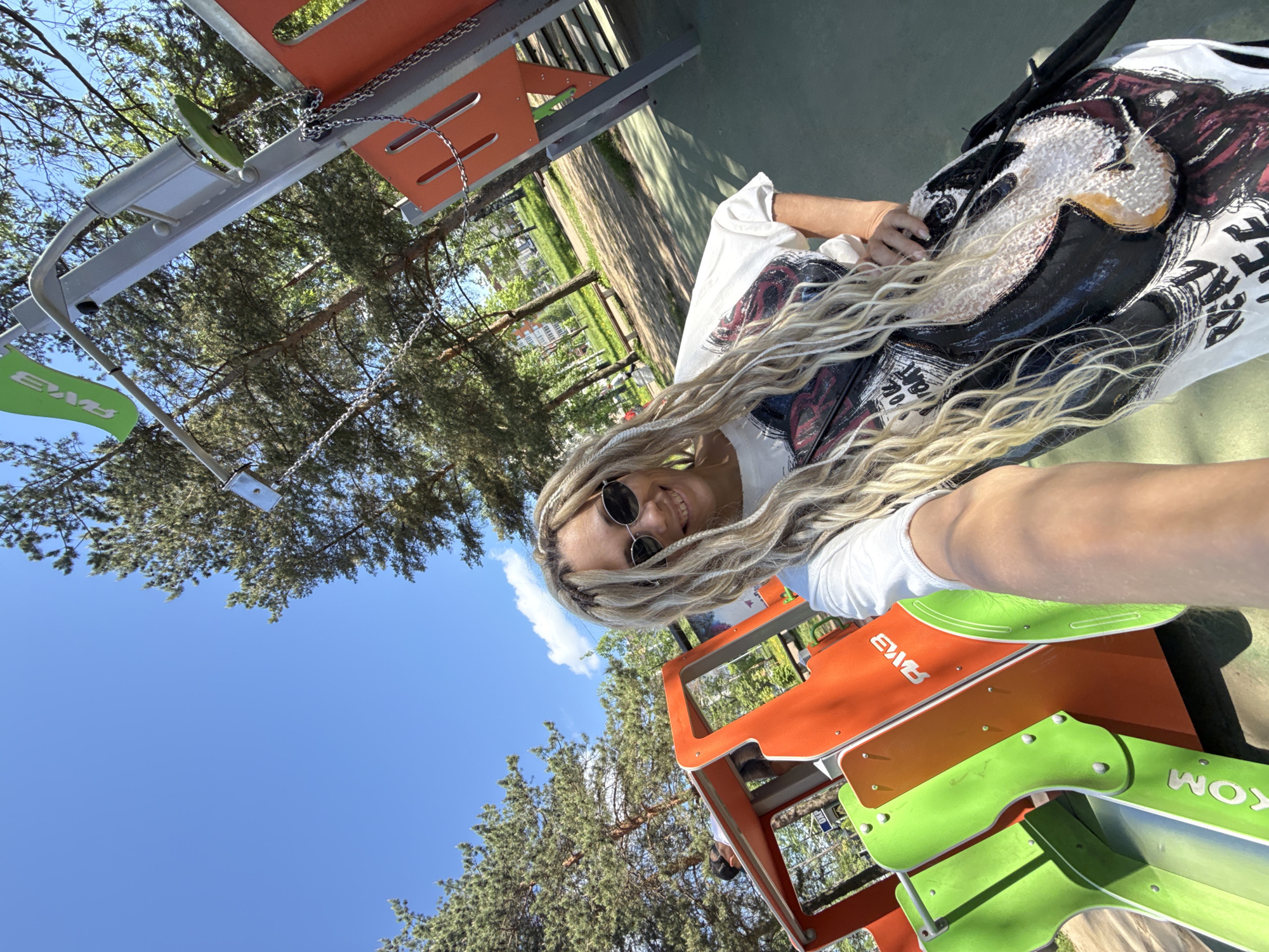 A woman with long, light-colored braids and sunglasses poses for a selfie at a playground in Unknown, Unknown. She is wearing a white t-shirt with a large graphic design. Various pieces of playground equipment, colored orange and green, are visible around her. The sky is blue with some clouds, and trees are in the background.