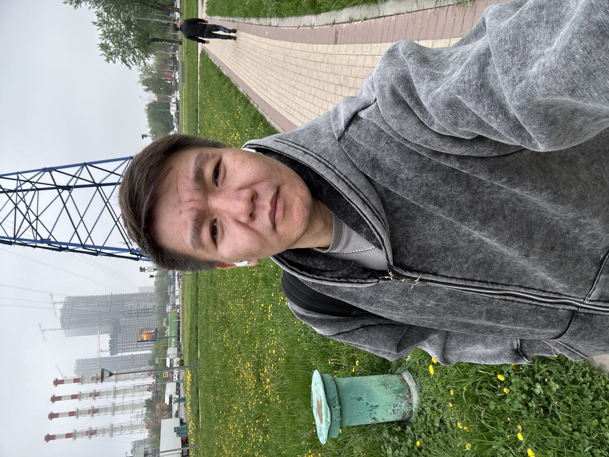 A rotated eye-level outdoor selfie shows a man wearing a gray hoodie, with a power line structure visible behind him on the left. In the background, there is a cityscape including tall buildings under construction with cranes, along with smoke stacks, and traffic on a street. The man is standing in an area with green grass and yellow dandelions. A green utility box is near the man's lower right. In the background, on the top right, there is a walkway with another individual walking away. Based on the architectural style and urban landscape, the location is likely in Moscow, Russia.
