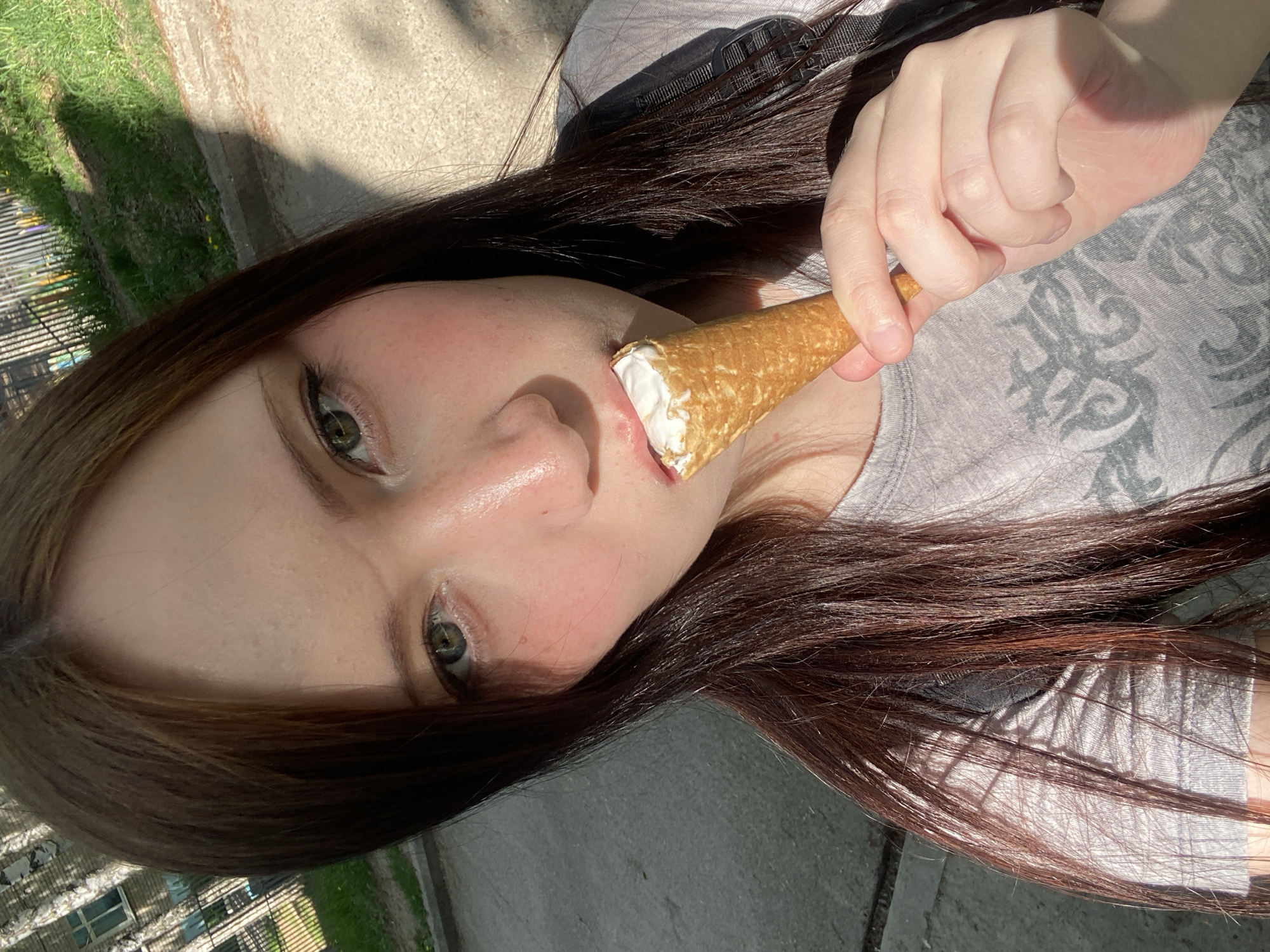 A person is shown in a close-up, angled shot, eating a cream-filled cone. The person is holding the cone with their right hand, placing it near their mouth. They have fair skin, dark hair, and green eyes. The person is wearing a gray shirt. The background includes concrete and some greenery, suggesting an outdoor urban environment.