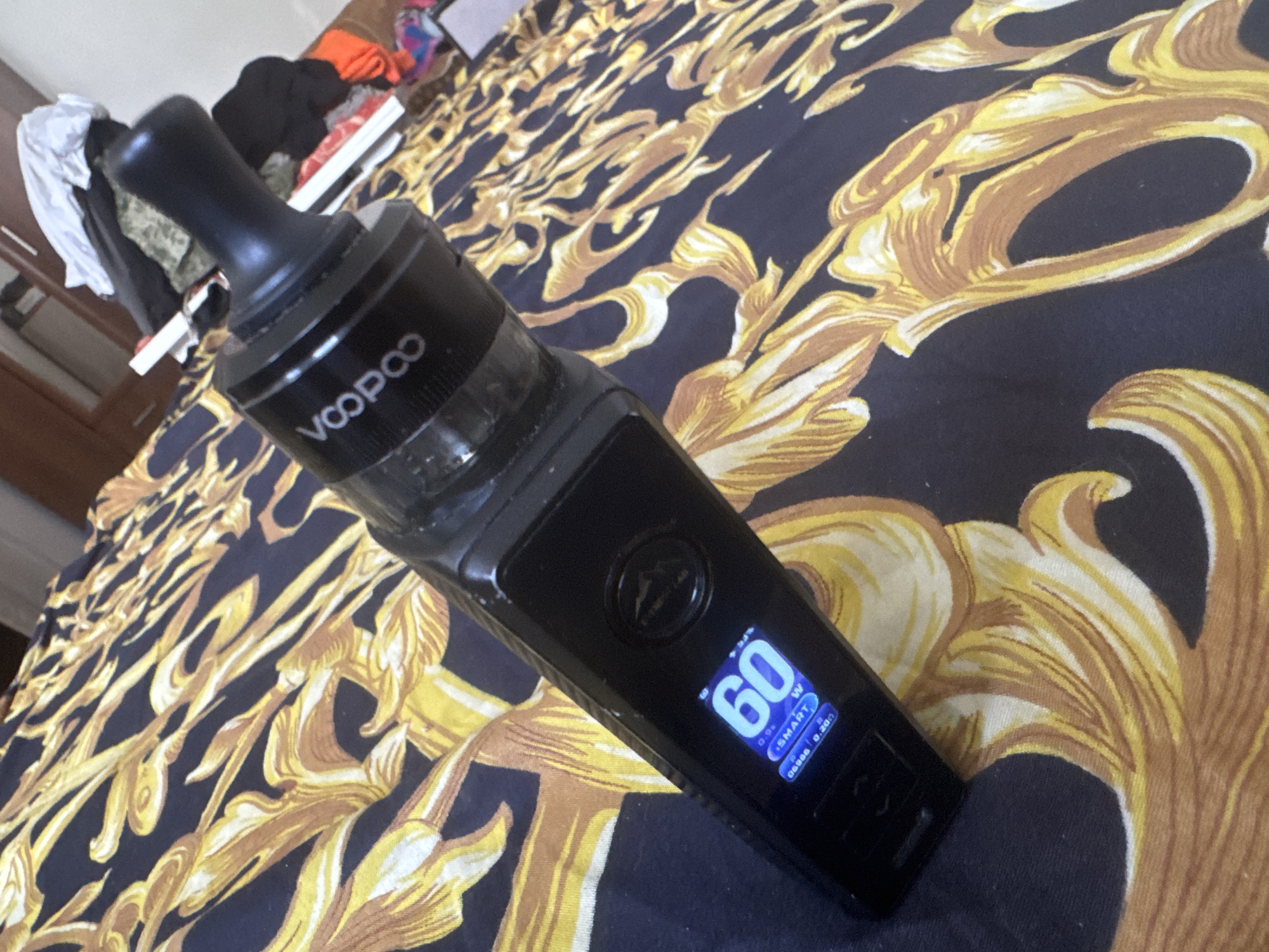 A black vape pen with a black mouthpiece rests on a dark blue bedspread with a gold baroque pattern. The vape pen's screen displays "60W" in large blue font, indicating the power setting. Below this, "SMART" is displayed in smaller white font, along with other readings. In the background, a closet with some clothes is visible. This image was taken indoors in a residential setting, likely in a bedroom.