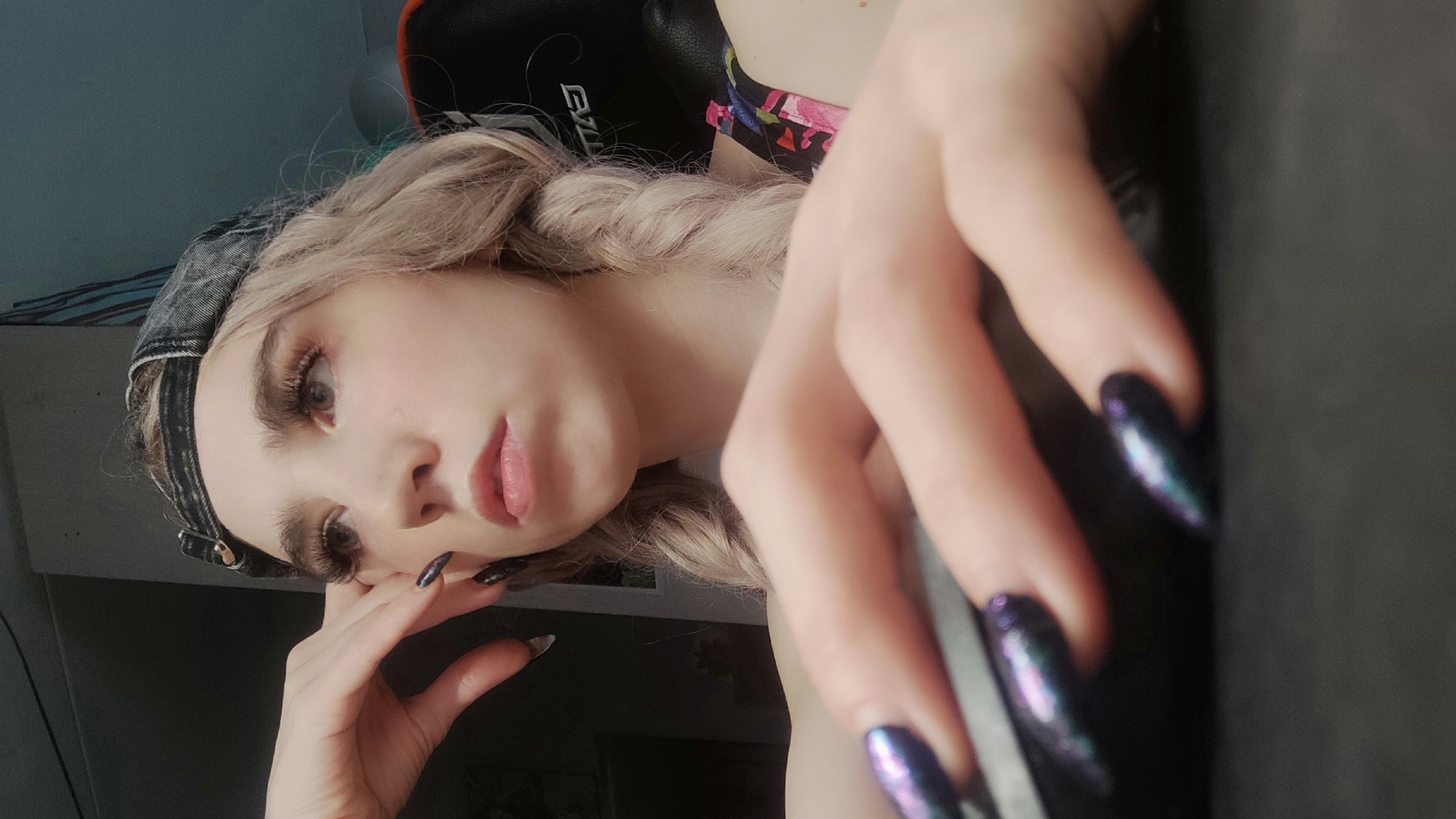 This close-up, angled portrait features a young individual, likely female, with light blonde hair styled in a braid. She wears a dark, denim-like cap and a top with a floral pattern, partially visible on her shoulder. Her face is prominent, showcasing defined eyebrows, long false eyelashes, and softly tinted lips. Her gaze is directed towards the camera, conveying a neutral or slightly contemplative expression.
Her hands are significant elements in the composition. Her left hand rests gently near her chin, while her right hand is positioned in the foreground, holding or resting on a large, dark, flat surface that extends across the right side of the image. Both hands feature long, almond-shaped fingernails adorned with a striking iridescent polish that displays shifts of dark purple, blue, and green hues.
The setting is an indoor room, possibly a bedroom or personal study. Behind the subject's head, part of a dark gaming chair with an orange accent is visible, and the letters "EZ..." can be faintly discerned on its headrest. The background is somewhat blurred, revealing a light-colored wall, potentially blue or grey. The lighting suggests it is daytime. The scene captures a casual moment in an indoor environment located in Chaikovsky, Russia.