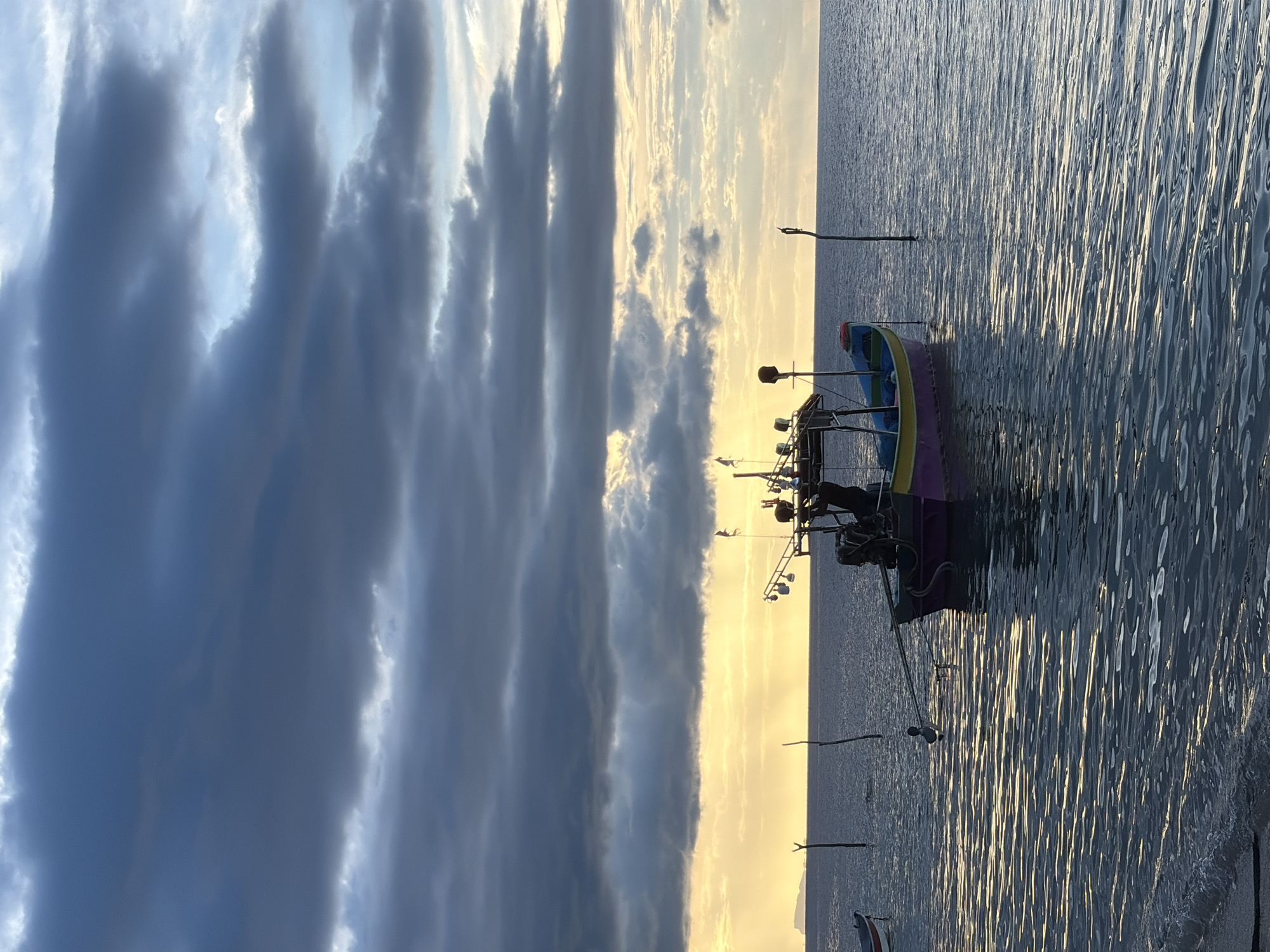 The image depicts a serene waterscape with a dramatic sky at sunset. In the foreground, a small, colorful boat, predominantly purple and yellow, is moored. It appears to be a fishing boat, with several fishing rods visible. The boat is gently rocking on the water, which reflects the warm hues of the setting sun, creating shimmering patterns on its surface.
The sky is a striking composition of dark, brooding clouds on the left, transitioning to a bright, golden glow on the right, characteristic of a sunset. This contrast suggests a dynamic weather pattern or the final moments of daylight. The overall scene evokes a sense of tranquility and the end of a day's work at sea.
The location is identified as Baan Thong Po, Thailand. The time of day is clearly sunset, with the low-angled sunlight casting a warm illumination across the water and sky. The weather appears calm, with the water showing only gentle ripples. The scene is peaceful and picturesque. No text is visible in the image.