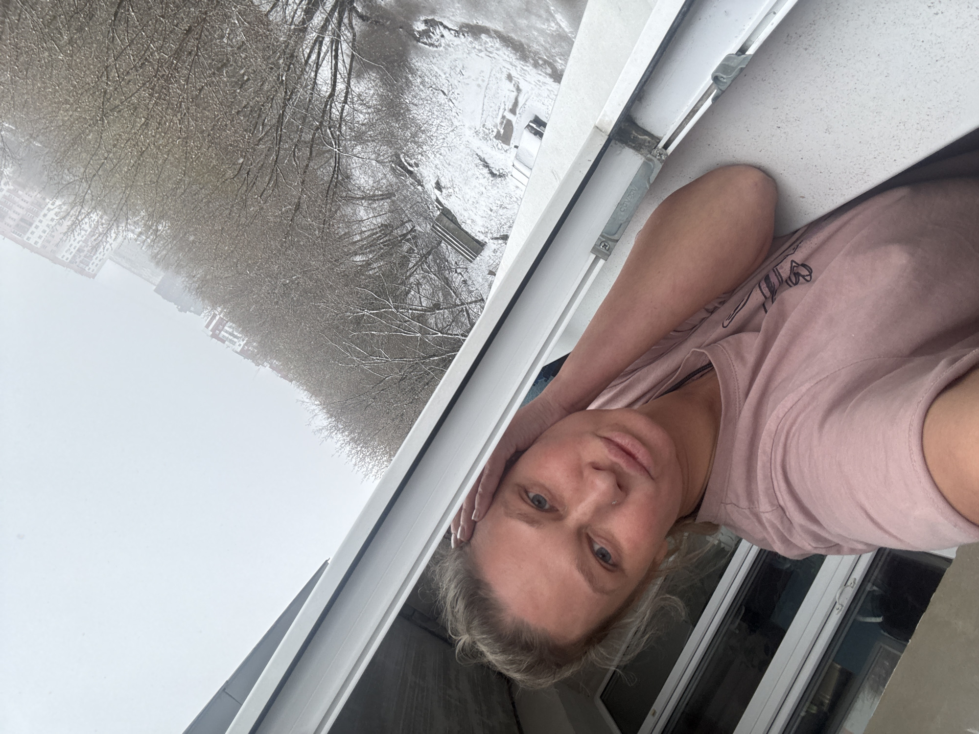 A fair-skinned woman with light blonde hair is captured in a vertical portrait, looking directly at the camera while leaning against a white window frame. She appears to be in her 30s or 40s and wears a light pink short-sleeved t-shirt with a subtle graphic design partially visible on the chest. A small stud piercing is visible on her left nostril. Her left hand is casually resting against the window sill, supporting her head, while her right arm is also visible in the shot.
The setting is an elevated view from inside a building, likely an apartment in Perm, Russia, looking out onto a snowy winter landscape. The window frames a scene dominated by bare trees heavily laden with snow and ice, suggesting ongoing or recent snowfall. In the distance, several multi-story residential buildings are visible through the wintry haze. The ground outside is completely covered in a thick blanket of snow.
The weather is clearly cold and snowy, with an overcast sky creating soft, diffused daylight. The activity involves the woman observing the winter scene or posing for a photo. A metal bracket within the white PVC window frame shows partially visible text that reads "COV". The overall mood is calm and contemplative, reflecting a quiet moment amidst a harsh winter day.