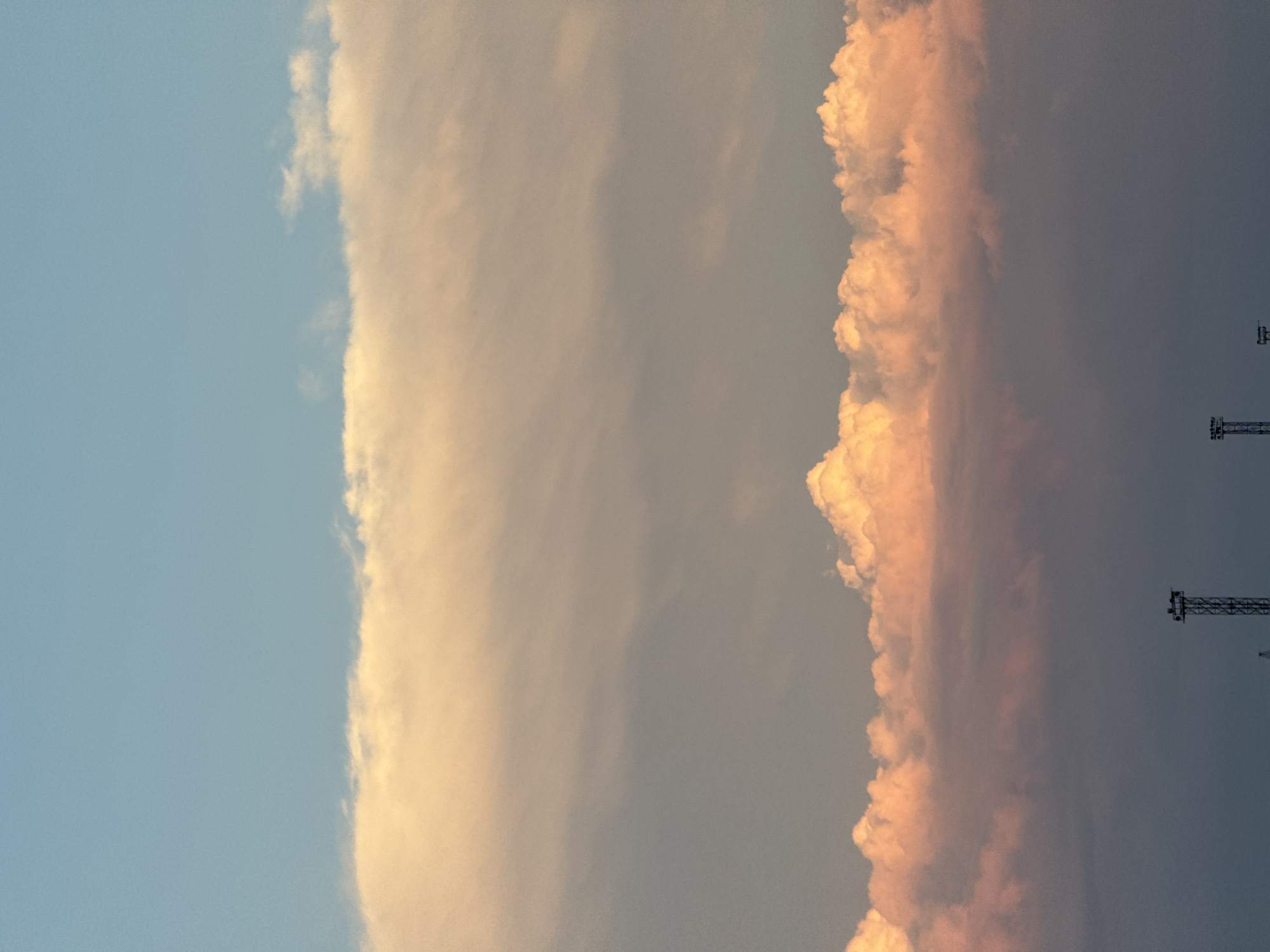 The image depicts a landscape under a pale blue sky with scattered white clouds. A broad band of lighter clouds, tinged with yellow and pink hues, stretches across the mid-section of the frame. Below this, a layer of darker, more subdued clouds fills the lower portion of the sky. In the distance, silhouetted against the dimming sky, are the tops of two industrial structures, possibly communication towers or cranes, suggesting an industrial or port area. The scene is likely captured during twilight, given the soft lighting and the warm tones of the clouds. The location is identified as Қоянды ауылдық округі, Kazakhstan, which implies a rural or semi-rural setting, though the presence of industrial structures could indicate proximity to a developed area or transportation hub. No people or specific actions are visible.