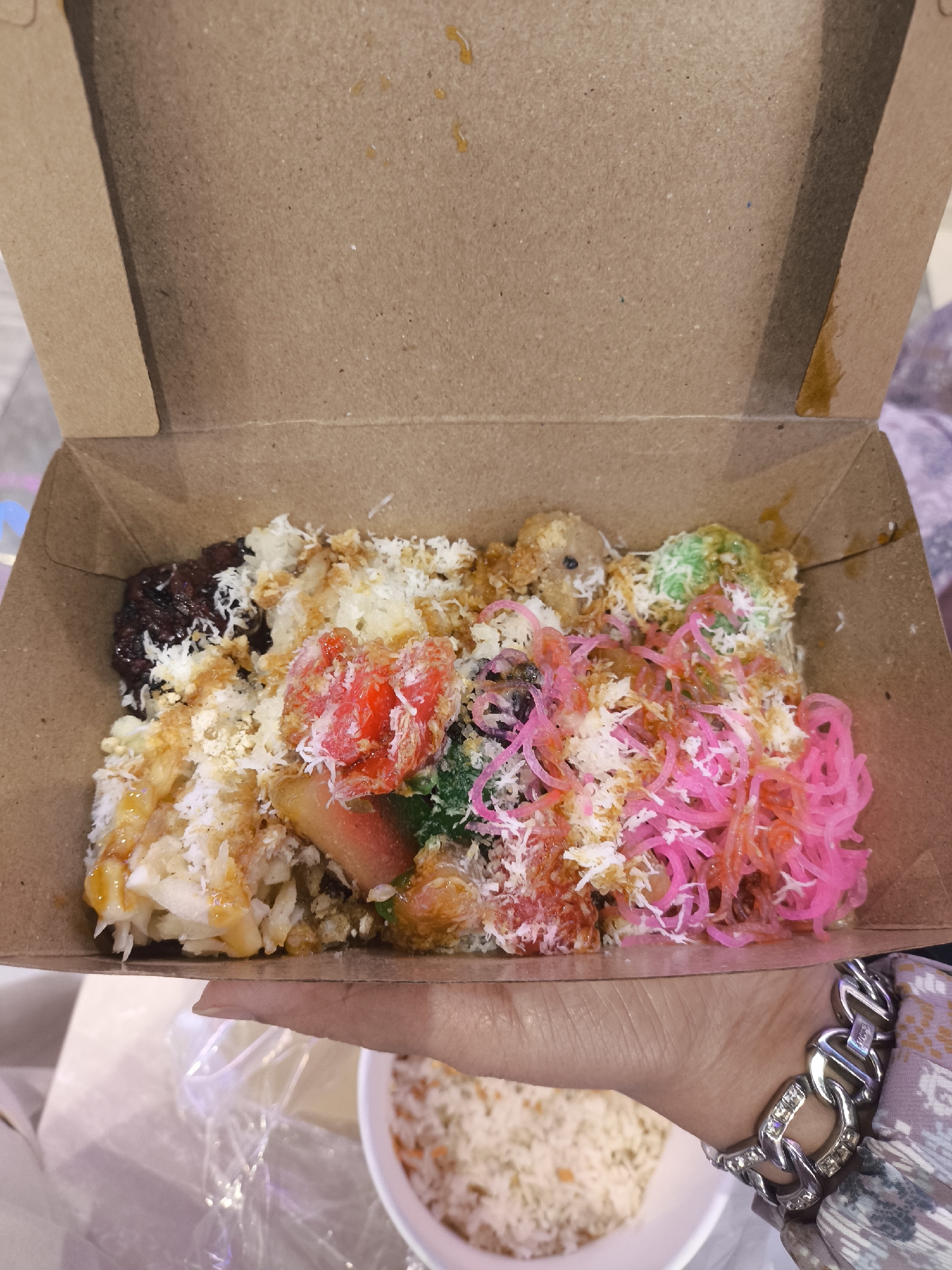 In Grogol, Indonesia, a person's hand, adorned with a silver bracelet, holds open a cardboard takeaway box filled with a colorful assortment of food. The box contains a base of white rice, topped with shredded coconut, small red and pink strands that appear to be noodles or candy, a few green globular items, and what looks like dark, sticky rice on the left. A drizzle of golden sauce is visible over the top layers of the food. The background suggests an indoor setting, possibly a food stall or market, with the bottom of the image showing a white bowl containing more rice and other ingredients.