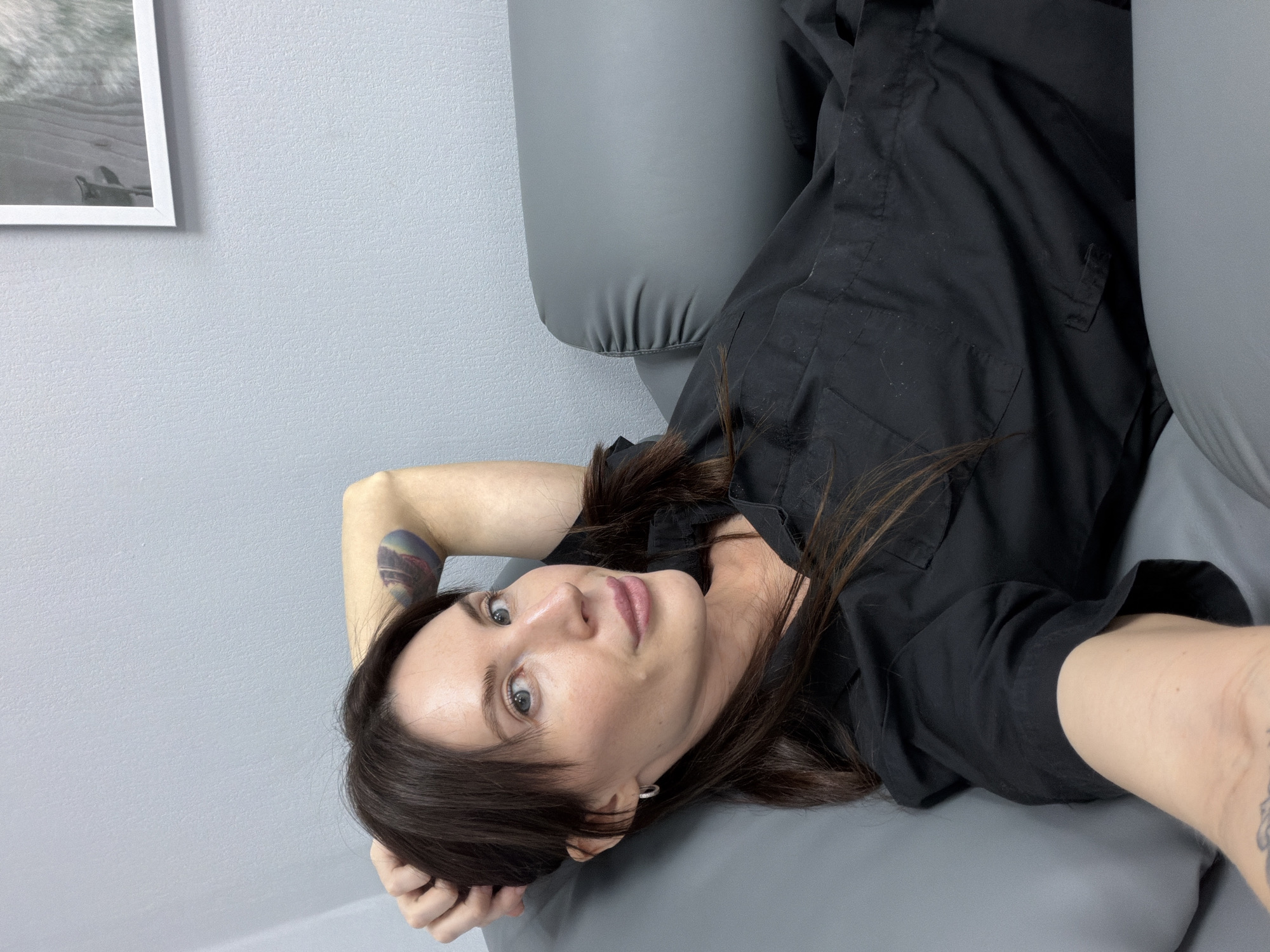 A woman lies on a gray couch in an indoor environment. She has fair skin, brown hair, and blue eyes, and she is wearing a black shirt. She has a tattoo on her arm. A framed picture hangs on the light gray wall behind her. The location appears to be in a city. The city is Perm, Russia.