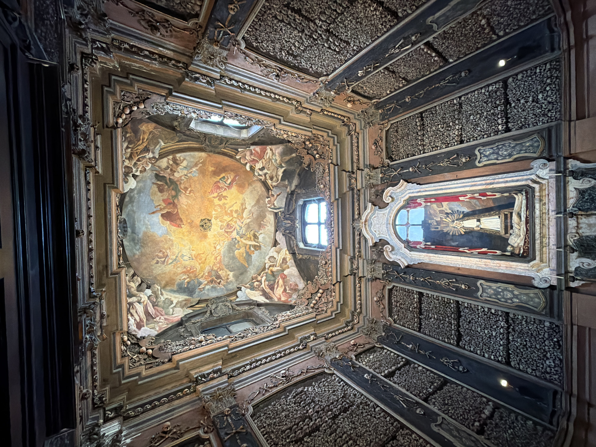 The image depicts the interior of the San Bernardino alle Ossa church in Milan, Italy, specifically the ossuary. The perspective is from below, looking up at the ceiling. The ceiling features an ornate painting with religious figures and a celestial scene. Walls are decorated with human bones, arranged in patterns and motifs. The ossuary also has decorative elements, including columns and frames. A statue of Madonna and child is visible behind a window.