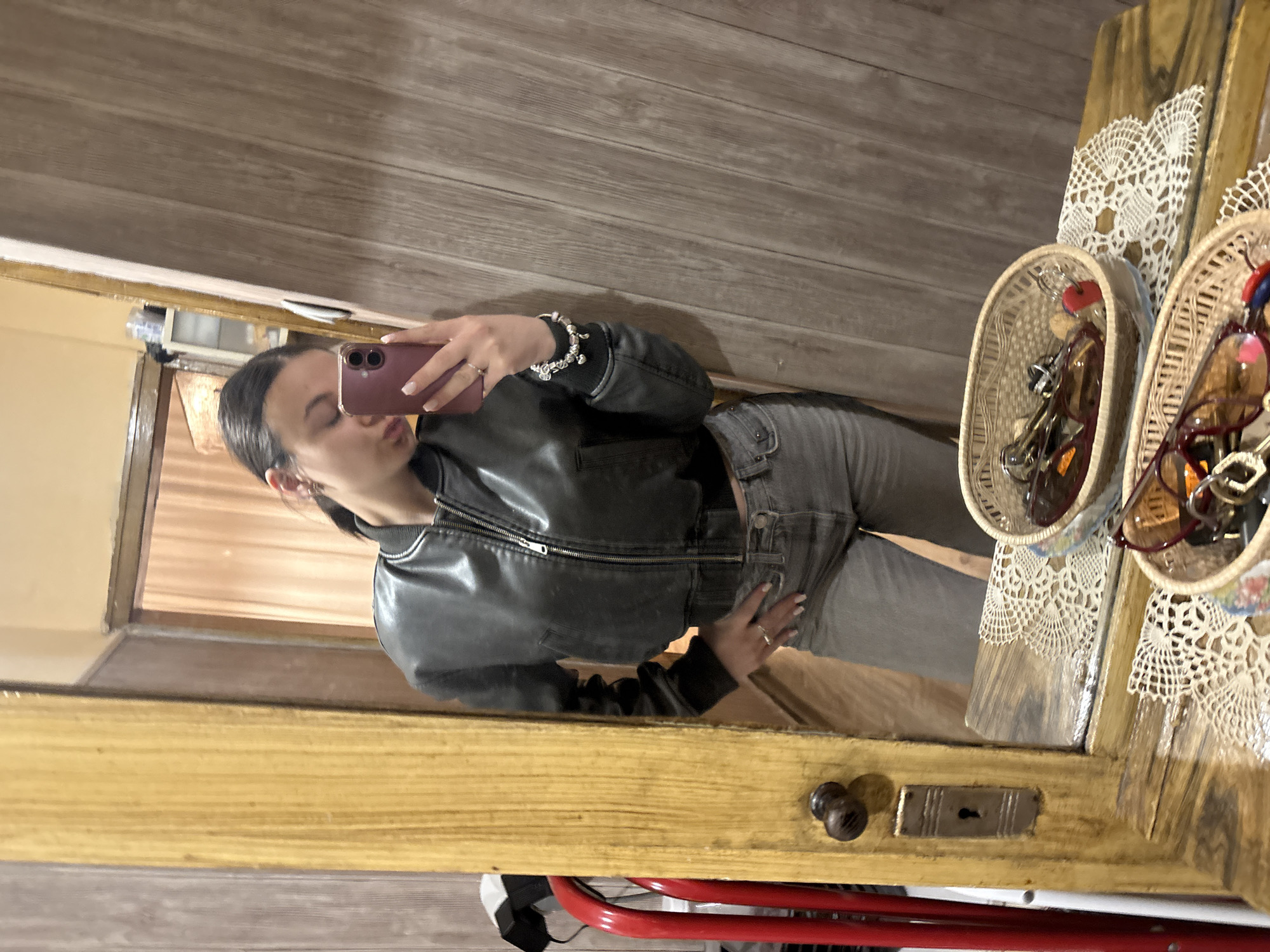 A young woman is taking a selfie in a mirror. She is wearing a black leather jacket and grey jeans. She is holding a pink phone in her right hand and has a silver bracelet on her wrist. The mirror is attached to a wooden cabinet or door that has a vintage style. There are two wicker baskets on top of the cabinet, filled with sunglasses. The environment appears to be indoors, possibly a shop or a home with rustic decor. The image does not provide enough information to confirm the location in Radomir, Bulgaria.