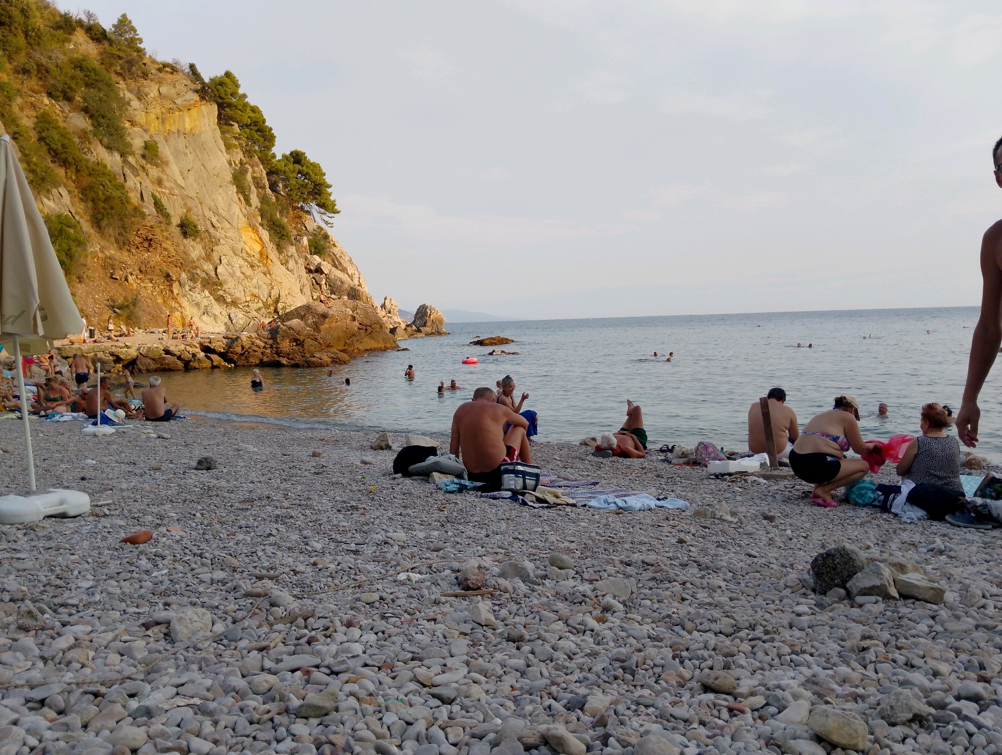 The image depicts a crowded pebble beach in Sutomore, Montenegro, during the late afternoon or early evening. A steep, rocky cliff covered in sparse vegetation dominates the left side of the frame, casting a shadow over a section of the beach. Several people are visible on the beach, some sunbathing, others sitting and talking, and a few are in the shallow water.
In the foreground, a large white beach umbrella base is partially visible on the left. The pebble-strewn ground is scattered with various personal items, including bags, towels, and clothing.
Several individuals are seated on towels or directly on the pebbles. To the right of the center, a man is seated with his back to the camera, appearing to be resting. Further to the right, a woman in a bikini is crouching, tending to some belongings, while another woman sits beside her. In the water, multiple people are swimming or wading, with a pink inflatable ring visible in the distance. The sea is calm, with a hazy sky overhead. The overall impression is one of a relaxed, popular beach scene in a coastal town.