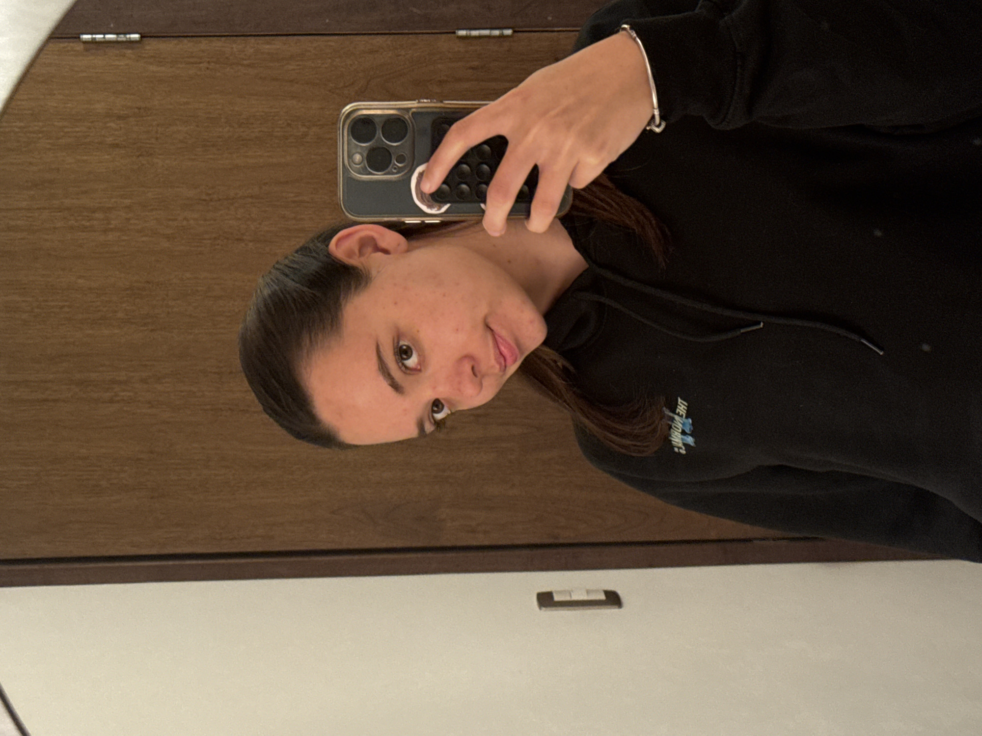 A person with light skin and brown hair, wearing a black hoodie and a silver bracelet on their left wrist, is taking a selfie in front of a mirror. They are holding a smartphone with a large camera array, likely an iPhone, in their right hand. The background shows a wooden door or panel above a white surface, possibly a wall or countertop. There is an electrical outlet below the wooden surface. The subject's face is turned slightly to the side, and their expression is neutral. There is no visible information in the image to determine a precise location in San Bernardino Tlaxcalancingo, Mexico.