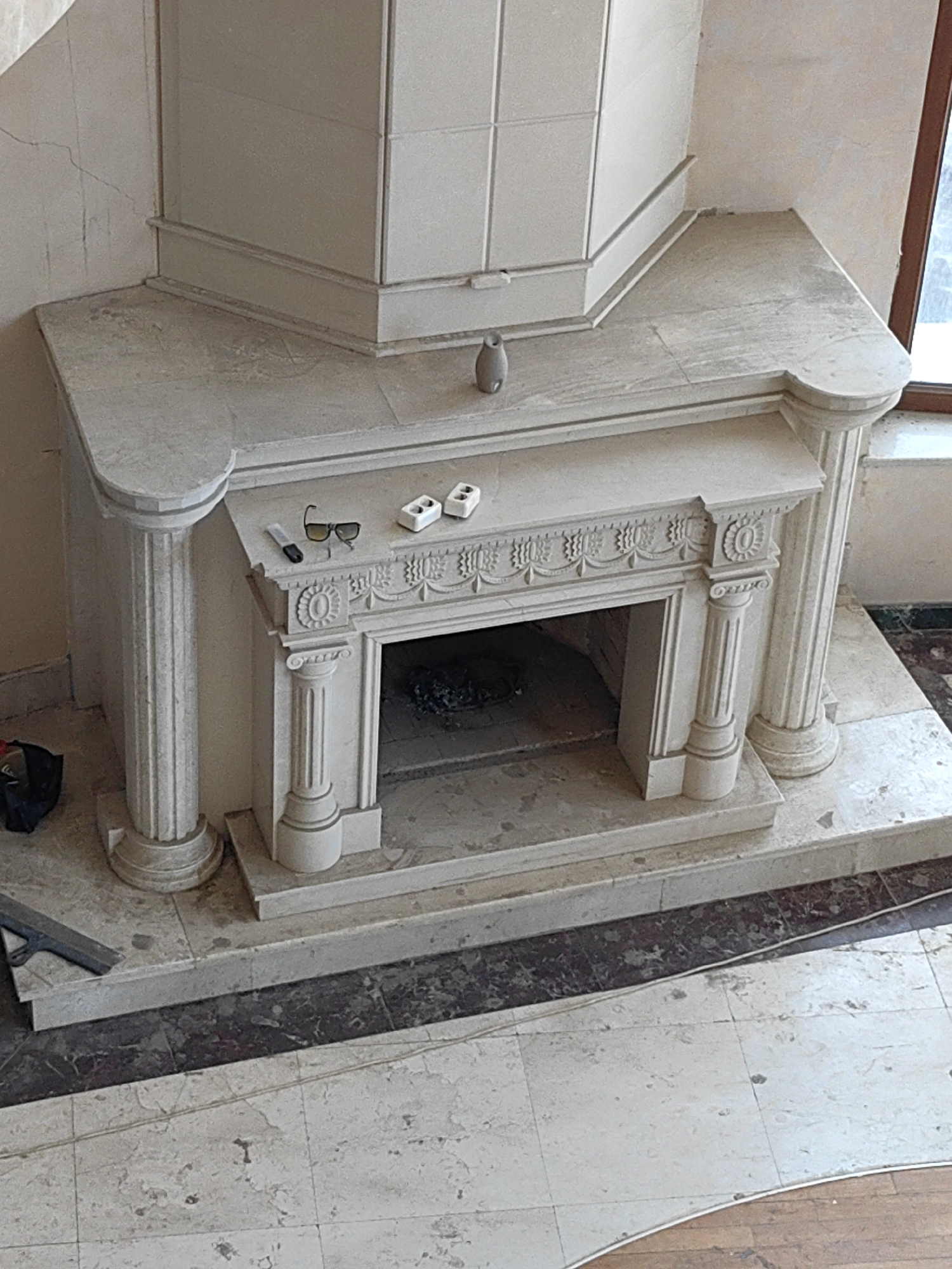The image shows a large, ornate marble fireplace in an unfinished state, likely during construction or renovation. The fireplace has a substantial mantelpiece supported by two large fluted columns on the sides and two smaller fluted columns flanking the firebox opening. Decorative carvings are visible on the frieze above the firebox. On the mantel, a pair of sunglasses, a small cylindrical object, and two white electrical outlets are placed. To the left of the fireplace, a black bag and a metal tool are visible on the floor. The surrounding walls appear to be plastered and have visible cracks. The floor is tiled with marble. The setting is indoors. There are no people visible in the image. The presence of construction materials suggests this is a residential or commercial space under renovation. Without additional context, the specific location within Shymkent, Kazakhstan, cannot be determined.