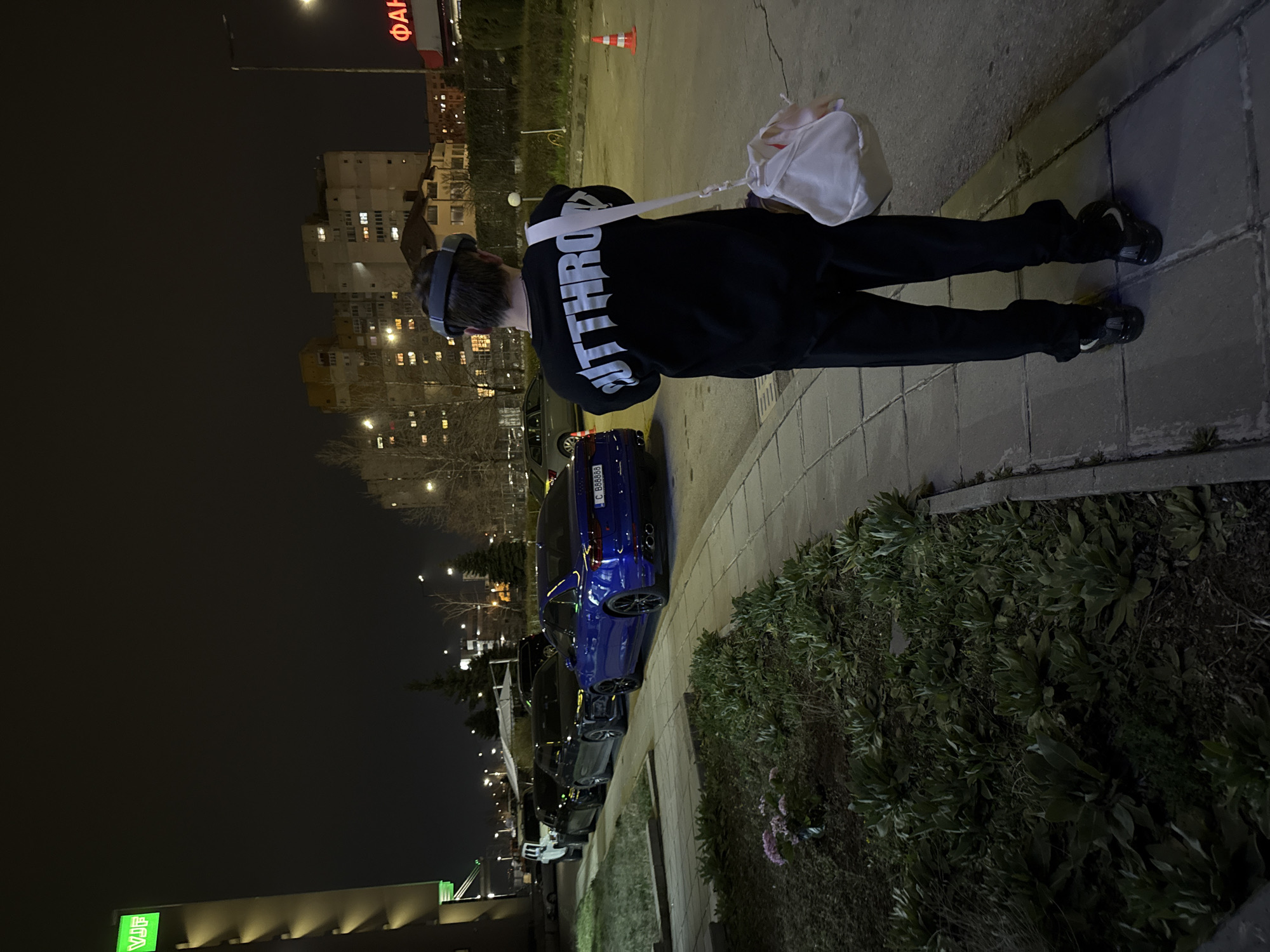 The image shows an individual wearing a black hoodie with the word "SITHROW" printed on the back, black pants, black shoes, and a VR headset. They are standing on a sidewalk next to a road, carrying a white bag.
A blue car with a license plate beginning with "C 8888" is parked next to the sidewalk. Other cars are parked further down the road.
The scene appears to be at night. The city is Sofia, Bulgaria, identified by the license plate style, and buildings in the background. A sign with red letters and the name "ФАМ" is visible in the distance. There is also a green sign with the letters "VIP." The environment includes a sidewalk, greenery, and street lights.