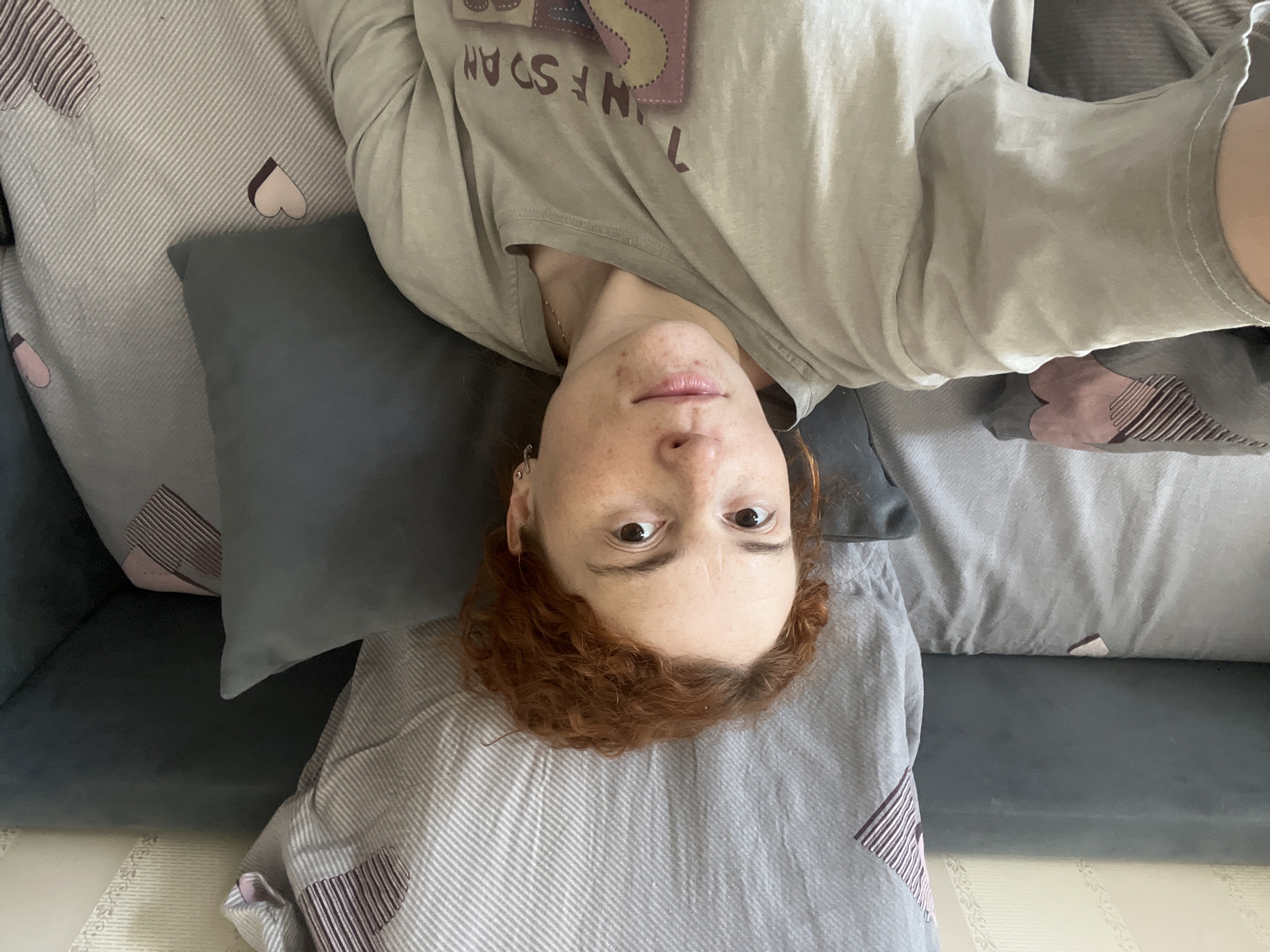 A woman with red curly hair and freckles is lying on a sofa covered with cushions and bedding. The sofa is dark gray, and the cushions and bedding are light gray with striped patterns and heart-shaped patches. The woman is wearing a gray t-shirt. The photograph appears to have been taken from an upside-down perspective. There is no evidence to confirm the location is Chaikovsky, Russia.