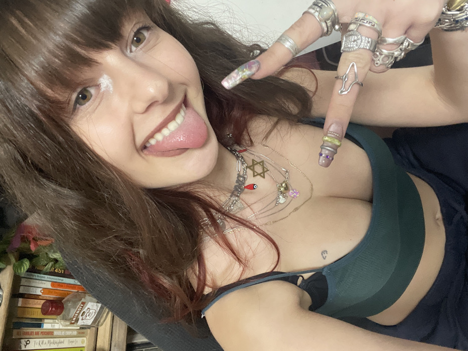 A young woman poses playfully, sticking out her tongue. She wears numerous rings on her fingers with long, decorated nails. Multiple layered necklaces adorn her chest above a dark green sports bra that reveals cleavage. A small heart tattoo is visible on her chest. To her left, a stack of books is partially seen.