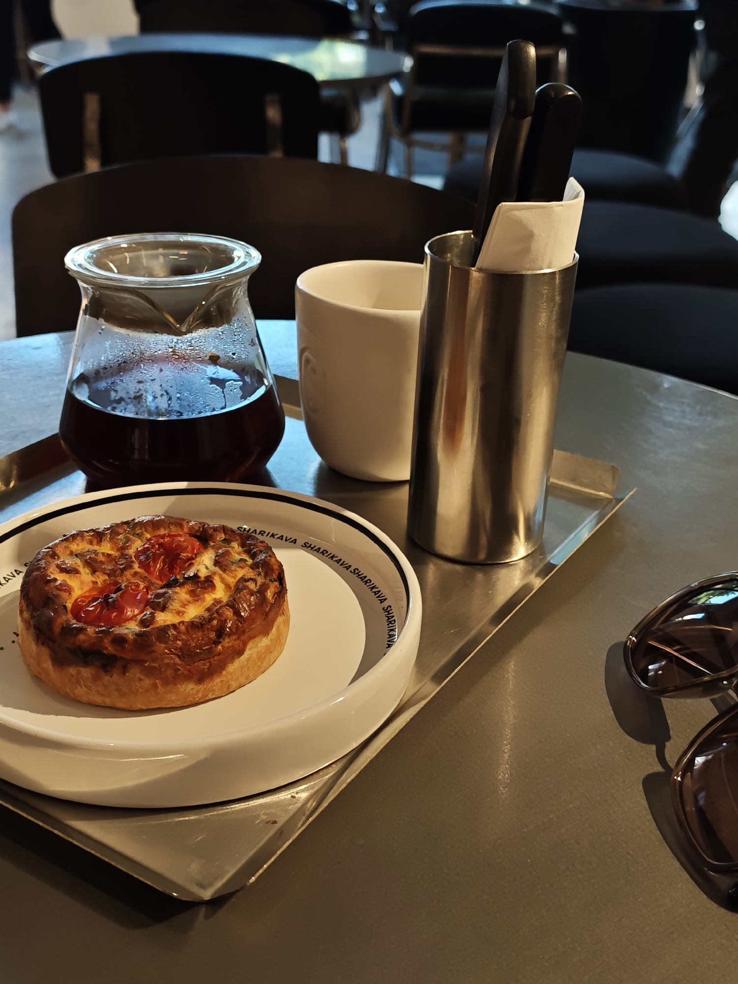 The image depicts a close-up view of a table setting at what appears to be a cafe or restaurant. On a metallic tray, there is a glass carafe filled with dark liquid, likely coffee, and a white mug. To the right of the mug is a stainless steel cylinder holding utensils and a white napkin. In the foreground, a round, golden-brown pastry topped with red cherry tomatoes and melted cheese is presented on a white plate. The plate has a black and white rim with repeated text. The background is blurred, showing dark chairs and tables, suggesting an indoor environment. The scene is lit by ambient light, creating reflections on the metallic surfaces and highlighting the textures of the food. The overall impression is one of a calm, possibly breakfast or coffee break setting. There is no discernible information in the image that would link it to Vinnytsia, Ukraine, nor are there any people or specific actions depicted.