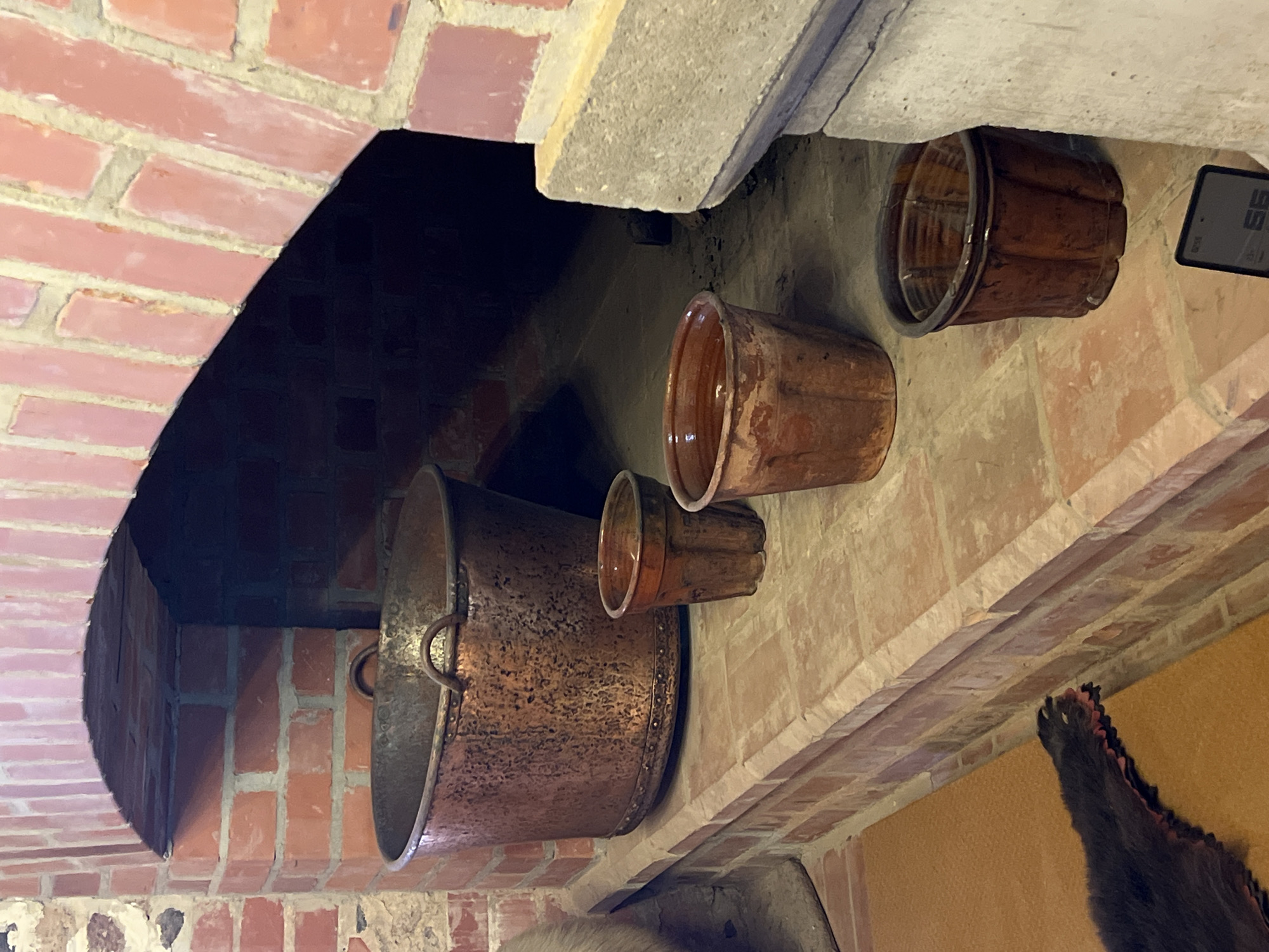 An interior shot shows a brick fireplace with an arched opening. On the hearth in front of the fireplace are four terracotta pots of varying sizes and one large copper basin. The largest pot, a copper basin, is in the center-left foreground. Three smaller terracotta pots are arranged to the right of the copper basin. A furry, dark-colored object with claws, possibly an animal pelt, is partially visible in the lower-left corner of the frame. A digital device displaying the number "56" is on the hearth in the lower-right corner. The scene is likely located in Trakai, Lithuania, given the historical and architectural context often found in the region.