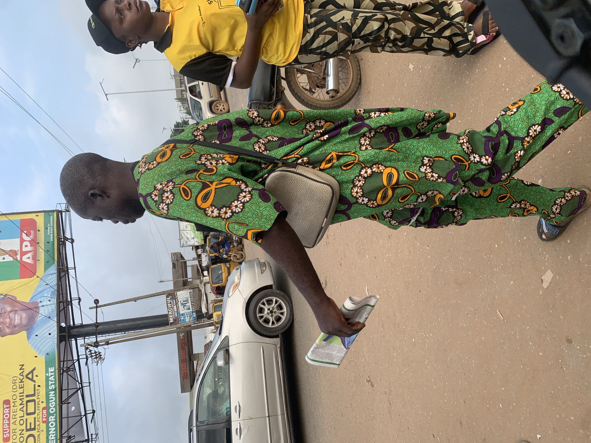 The image captures a street scene in Shagamu, Nigeria, likely near a busy intersection given the presence of vehicles and a billboard. In the foreground, a man walks towards the right, holding a folded newspaper in his left hand. He is wearing a vibrant green and purple patterned traditional outfit and a crossbody bag. To his right, a woman wearing a yellow polo shirt with black trim and a black baseball cap also looks towards the right. She is holding a handheld electronic device. Behind them, several vehicles are visible, including a silver minivan, a white SUV, a motorcycle, and some yellow tricycles, suggesting active traffic. A large billboard promoting "Governor, Ogun State" and "APC" is prominently displayed in the background, indicative of political campaigning. The ground is a dusty, unpaved road, and electrical poles and wires are visible overhead, typical of an urban Nigerian environment. The overall impression is a snapshot of daily life in a bustling Nigerian city.