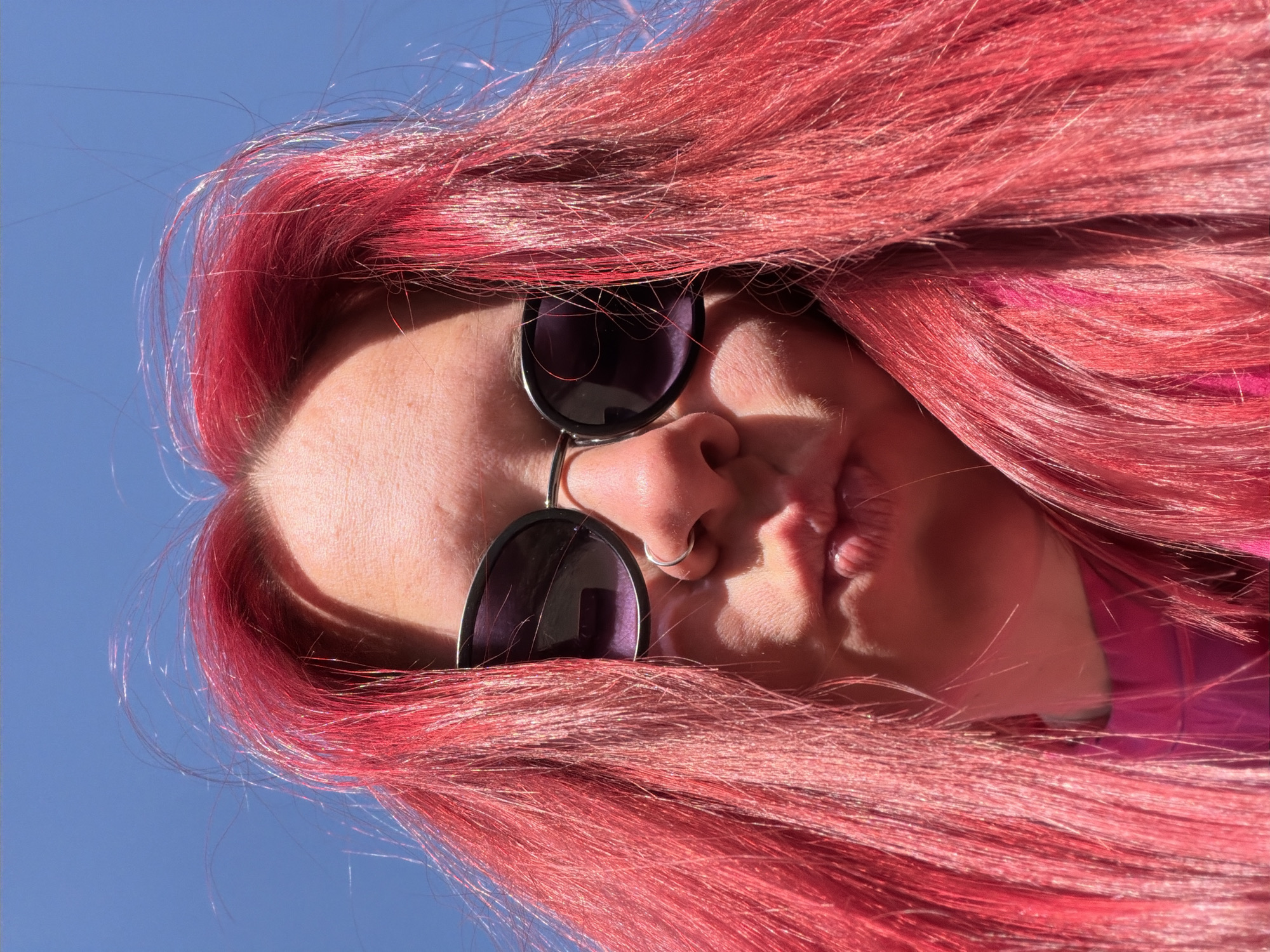 A close-up, tilted low-angle shot shows a person with red hair wearing sunglasses and a nose ring. The background is a clear blue sky. The person is pursing their lips. The location information is not possible to determine from the image.
