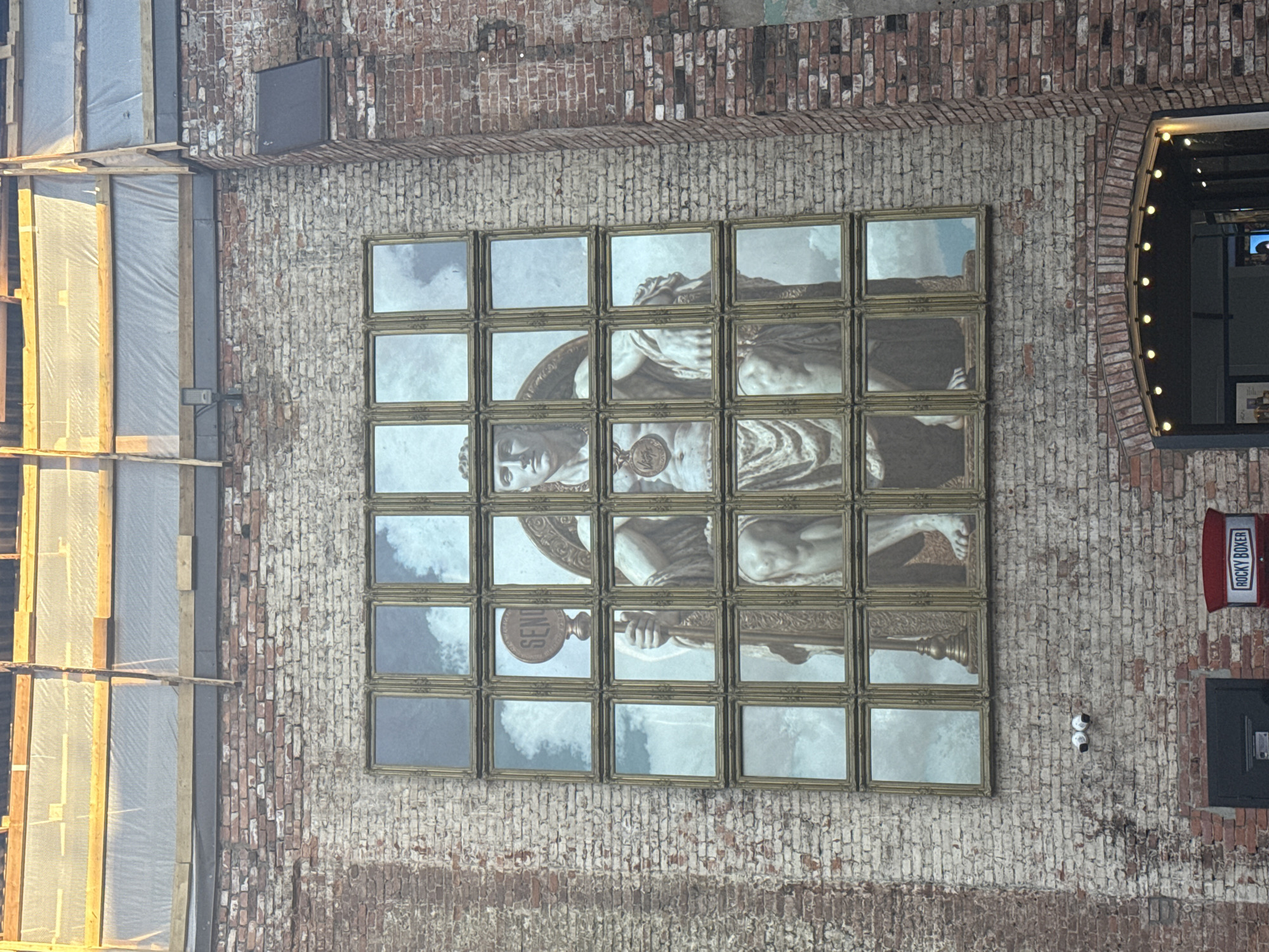 The image depicts a brick building exterior. A square grid of 20 framed pictures displaying a classical statue fragment is mounted on the brick wall. Adjacent to the grid is an arched doorway with lighting. To the right, there is a "ROCKY BOXER" sign over a door. Two security cameras are mounted between the artwork and the door. Scaffolding is visible on the left side of the image. The location is identified as Saint Petersburg, Russia.
