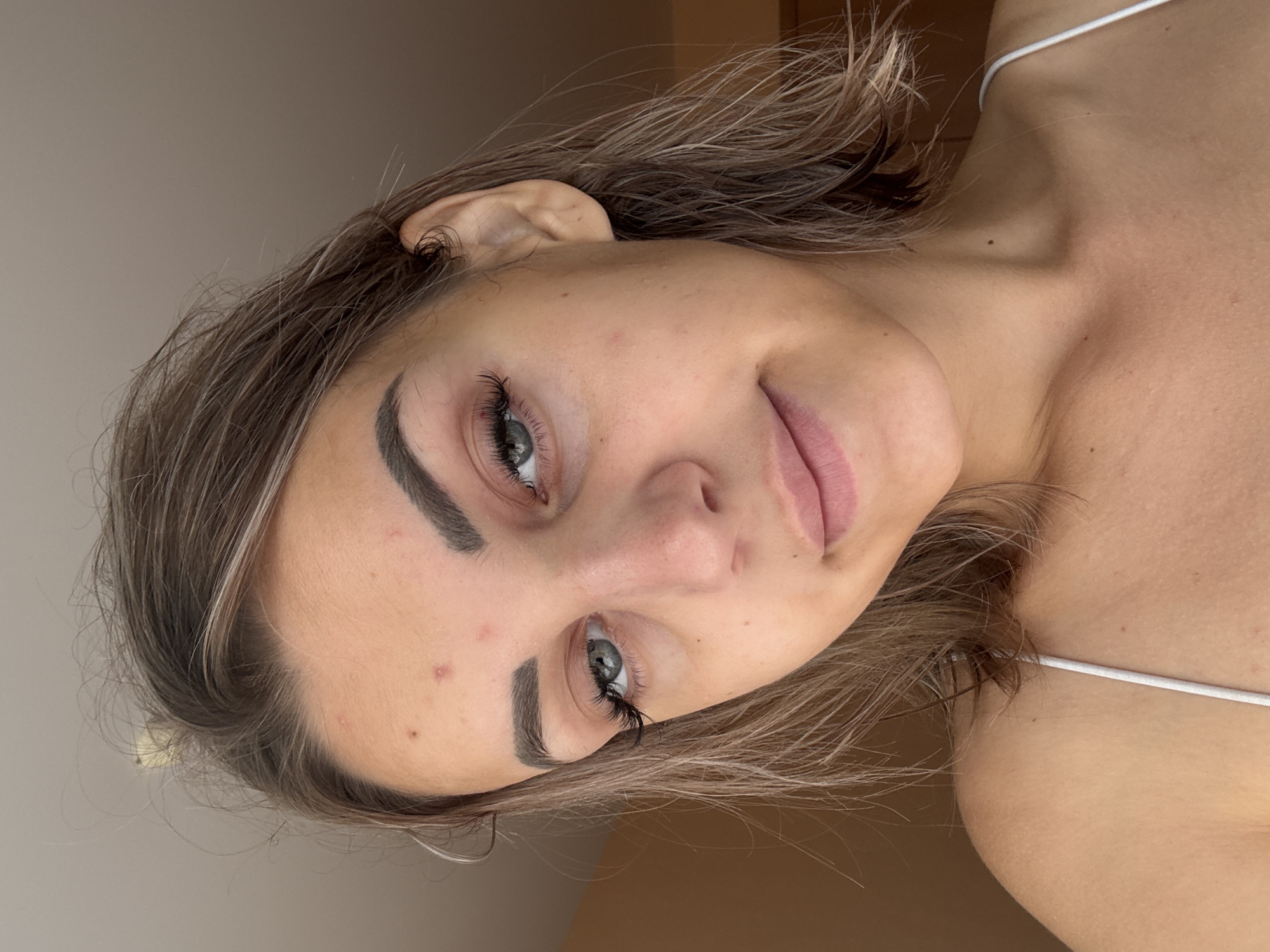 A close-up, head-and-shoulders studio portrait of a young woman with light brown hair and blue-green eyes, looking directly at the camera with a subtle smile. She has notable blemishes on her forehead and cheeks, and appears to be wearing false eyelashes. Her attire consists of a white camisole strap visible on her left shoulder. The background is a plain, light brown wall. The image was taken indoors, likely in a residential setting in Kholmsk, Russia, given the subject's nationality and the commonality of such domestic scenes.