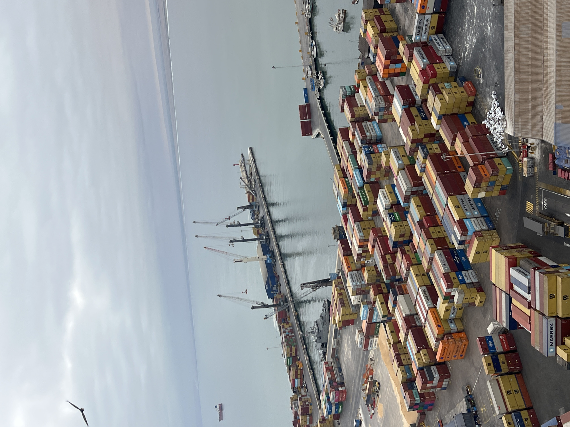 This detailed, high-angle image depicts the Port of Arica in Arica, Chile, under an overcast, light grey sky. A dark-plumaged bird, possibly a raptor or large seabird, is in flight with extended wings in the upper left quadrant of the sky.
The Pacific Ocean, calm and greyish-blue, extends from the mid-ground to a distant, hazy horizon line where it meets what appears to be a low-lying coastal landmass or atmospheric haze. A red-hulled commercial vessel is visible in the distant upper left.
The port infrastructure includes two piers. The larger, primary pier in the center-left features four prominent gantry or jib cranes, two of which have suspended elements and are positioned over the water. A large blue industrial building with "TPA" (Terminal Portuario Arica) clearly visible on its side is situated on this pier. Docked alongside this pier is a grey patrol vessel or military ship. To the right, a smaller, secondary pier extends into the water, equipped with a light or beacon at its end. Several smaller white-hulled fishing or recreational boats are moored at this secondary pier.
The lower two-thirds of the image are dominated by a sprawling container yard. Thousands of intermodal shipping containers, primarily branded "MAERSK," "MAERSK LINE," "CMA CGM," "MCC," and "ONE," are densely stacked in various configurations, typically two to five high. The containers display a diverse color palette, including red, blue, yellow, green, orange, white, and brown.
Within the container yard, excavation equipment is visible on a pile of granular material, likely sand or aggregate, in the mid-left. Various trucks and container handling vehicles are positioned among the container stacks. A large industrial building with a corrugated metal roof occupies the lower right foreground. A cluster of approximately ten individuals, appearing as small white figures, is visible on the ground near a stack of containers in the lower central-right. The word "PARE" (Spanish for "STOP") is painted in white on the asphalt in the lower left corner.