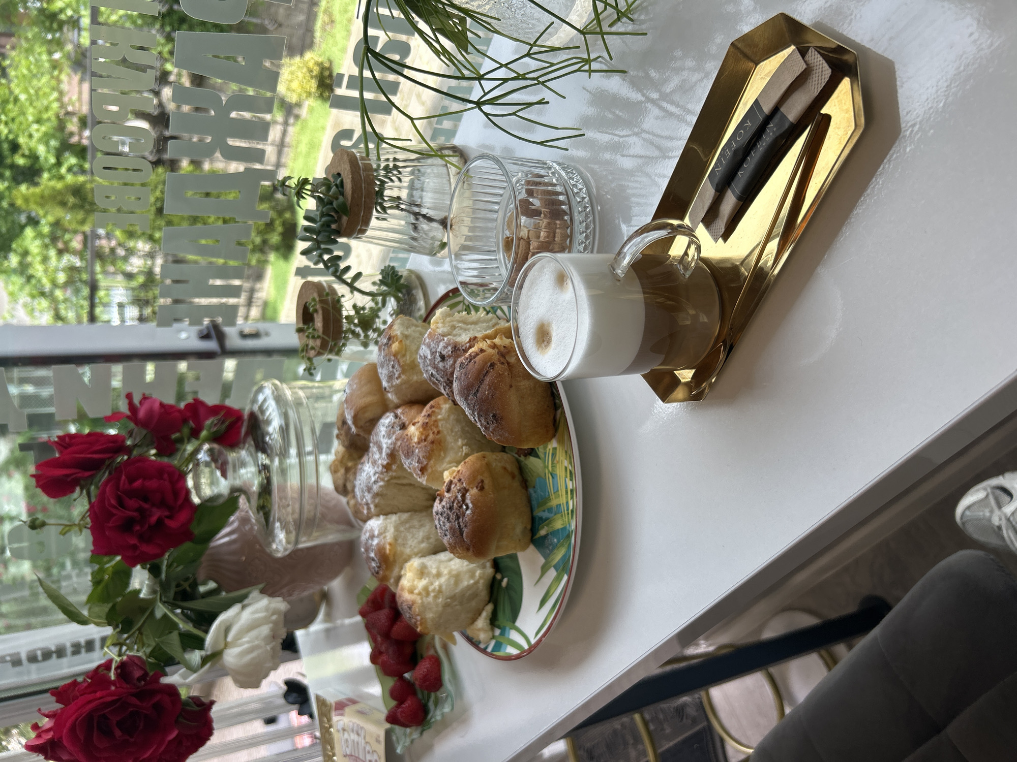 The image depicts a table setting in what appears to be a cafe or restaurant, situated in Unknown, Unknown, as indicated on sugar packets. A plate of scones is centrally placed, complemented by a glass of coffee and sugar packets on a golden tray. A vase of roses is present, alongside additional small plants and jars. The scene is shot from an elevated perspective, suggesting a bright, casual dining environment.
