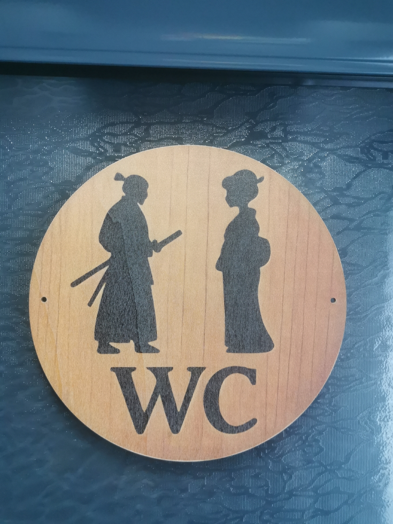 A circular wooden sign, indicating a restroom, is centrally displayed against a dark blue, textured background. The sign features two stylized black silhouettes: a male figure on the left, depicted as a samurai with a topknot and a sheathed sword, and a female figure on the right, dressed in a kimono with a traditional hairstyle. Both figures stand facing each other. Below the silhouettes, the bold, black letters "WC" are prominently visible, signifying "Water Closet." The wooden sign has a light brown hue, revealing its natural grain, and contains two small mounting holes on either side. The dark blue background surface has a glossy appearance and is adorned with a subtle, intricate relief pattern, possibly floral or organic, over a finer, grid-like texture. The scene implies an indoor setting where the sign directs visitors to facilities with a distinctive Japanese cultural theme. The image was captured in Blagoevgrad, Bulgaria.