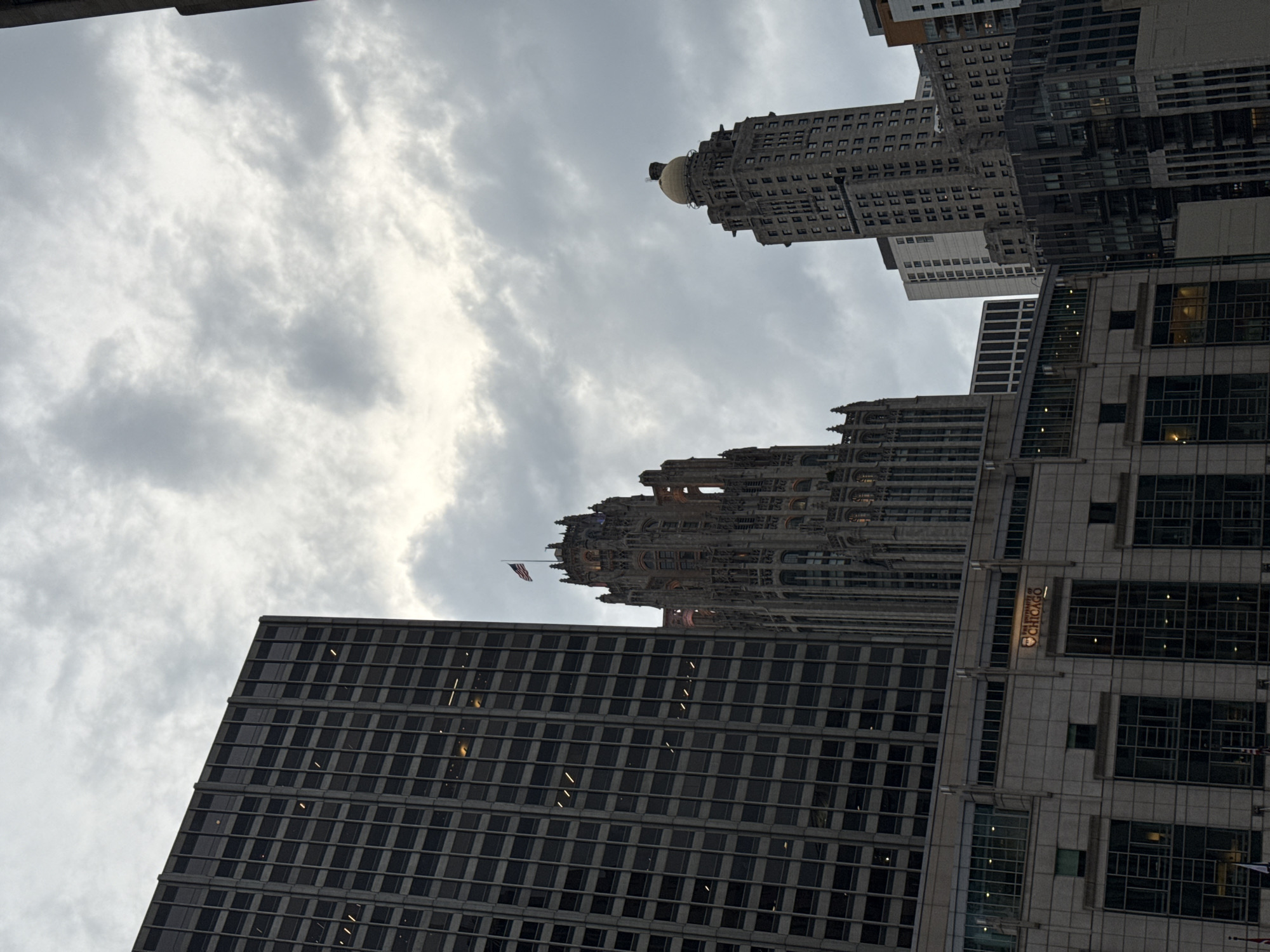 The image depicts a cluster of modern and historic skyscrapers in Chicago, Illinois, under a cloudy sky. In the foreground, a large, dark-colored, rectangular skyscraper dominates the left side of the frame, with its grid of windows stretching upwards. To its right, a Gothic-style skyscraper, illuminated from within, rises dramatically, topped by an American flag. Further to the right, another skyscraper features a distinctive golden dome. The lower portion of the image shows the facade of a building with the illuminated logo "THE UNIVERSITY OF CHICAGO" visible. The overall atmosphere is somewhat somber due to the overcast weather.