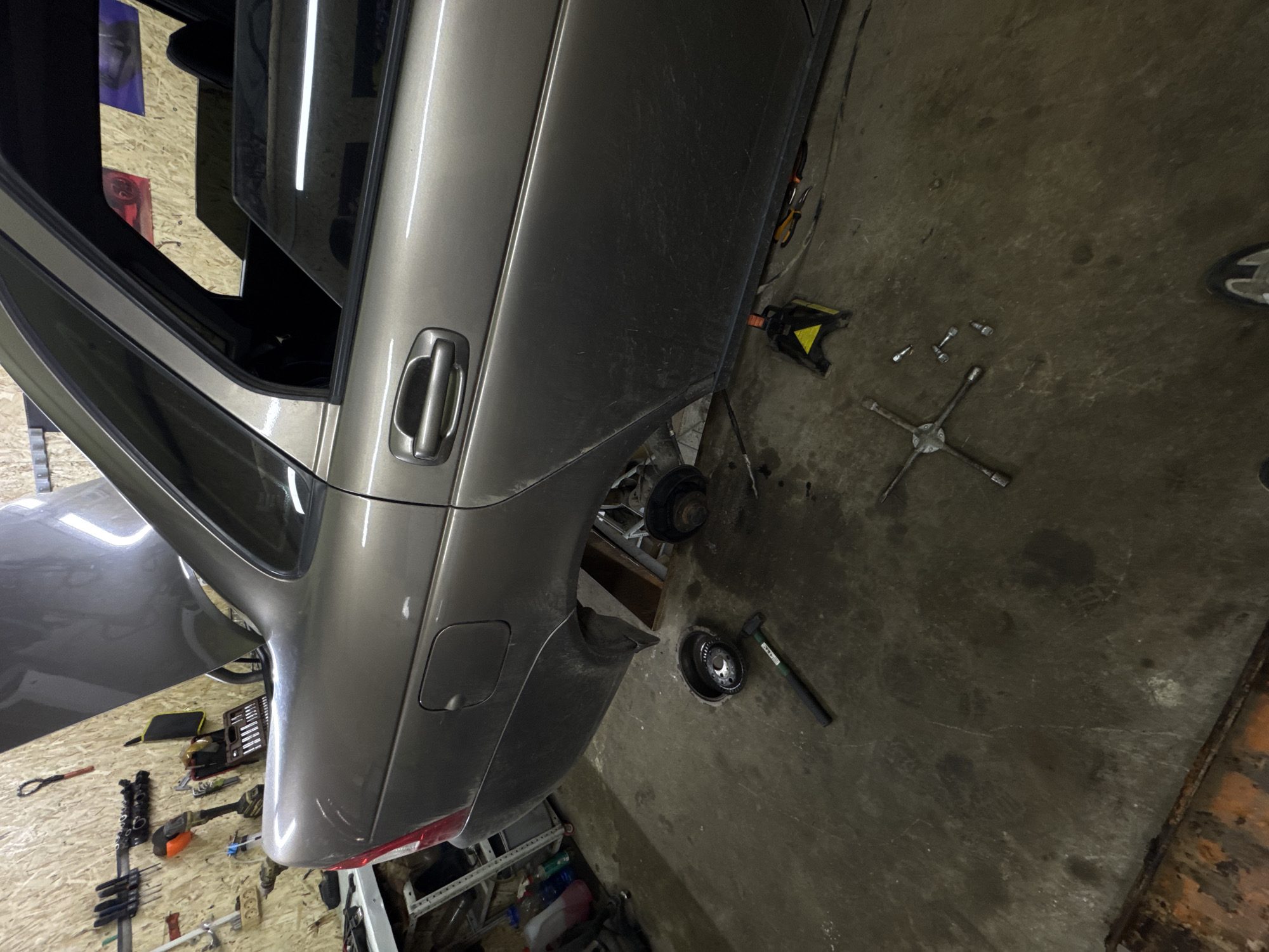 The scene depicts a dark metallic gray sedan undergoing repair or maintenance in an indoor garage or workshop, located in Perm, Russia. The vehicle's trunk is open, and both rear wheels have been removed. The car is elevated on at least one yellow and black jack stand, visible beneath the right rear passenger door sill.
On the vehicle's left rear, the wheel hub and exposed suspension components are visible. Directly below this area, on the concrete floor, a metal bucket-like container holds a complex mechanical component, possibly a gear assembly or bearing. A black hammer with a green handle rests near this container. On the right rear, the brake drum or rotor assembly is exposed.
The workshop floor is concrete, heavily stained with oil, grease, and dirt, consistent with a working environment. Visible on the floor near the vehicle's right side are a four-way lug wrench and several loose lug nuts. A person's right foot, wearing a white and grey sneaker, is partially visible in the lower right corner, indicating human presence.
The walls of the garage are clad with light-colored wooden panels (possibly plywood or OSB), serving as a pegboard for various tools. Hung on the wall are several wrenches, screwdrivers, a power drill, and a black tool case containing a socket set. Through the car's rear windows, reflections or posters are visible on the opposite wall, one appearing predominantly red and another blue/purple. A white shelving unit with bottles and containers is visible in the far-left background. The overall lighting suggests artificial illumination from overhead sources.