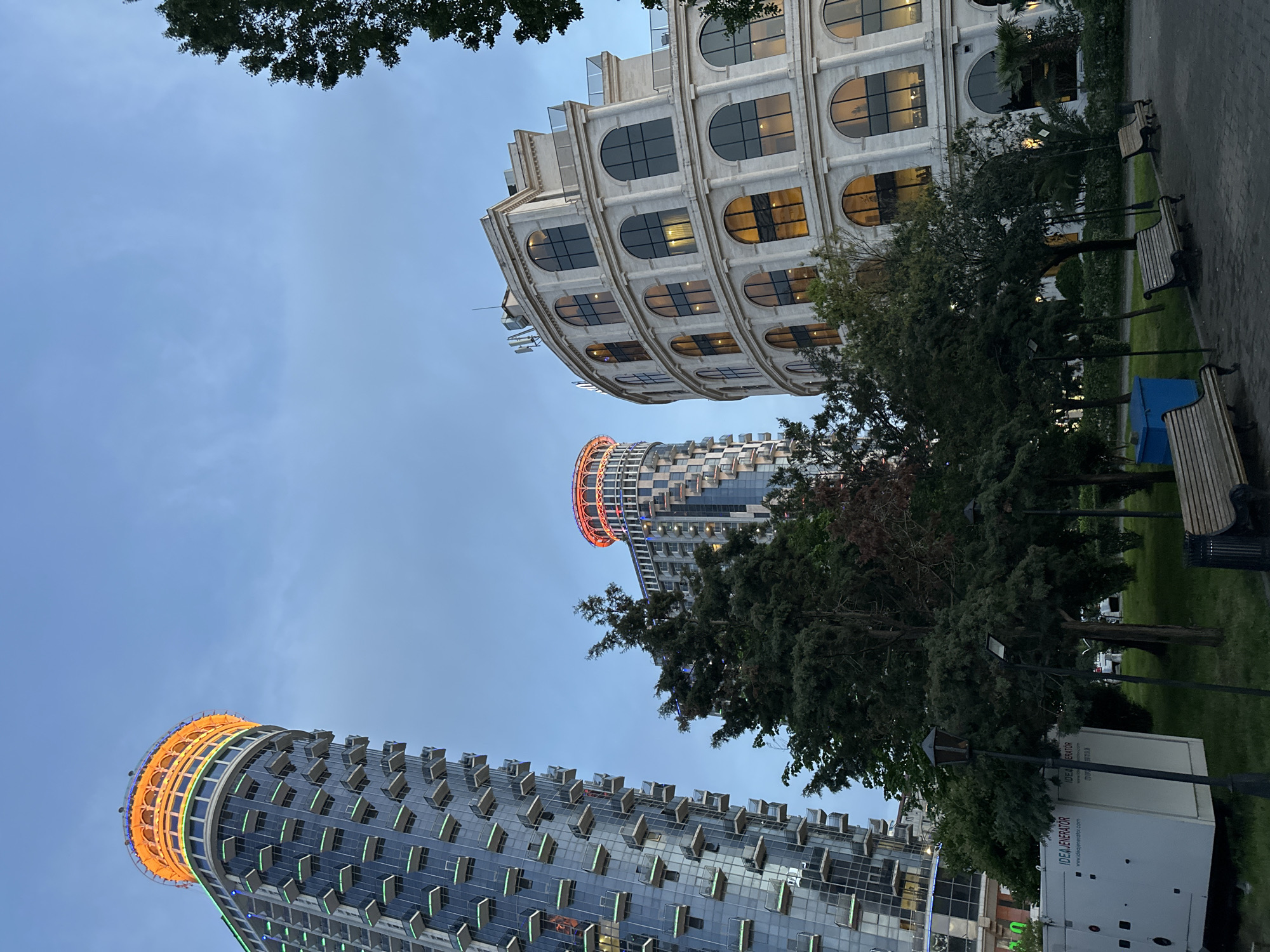 The image shows a view looking up at three prominent buildings in Batumi, Georgia, against a blue sky. The buildings are of different architectural styles. The closest building to the viewer, on the lower left, is a modern high-rise with rounded balconies. The top of this building is lit with orange lights. A second building, partially visible behind the first, has a cylindrical shape and a red-lit top. The third building is a white, multi-tiered structure with arched windows. In the foreground, there are trees, park benches, and a white generator trailer.