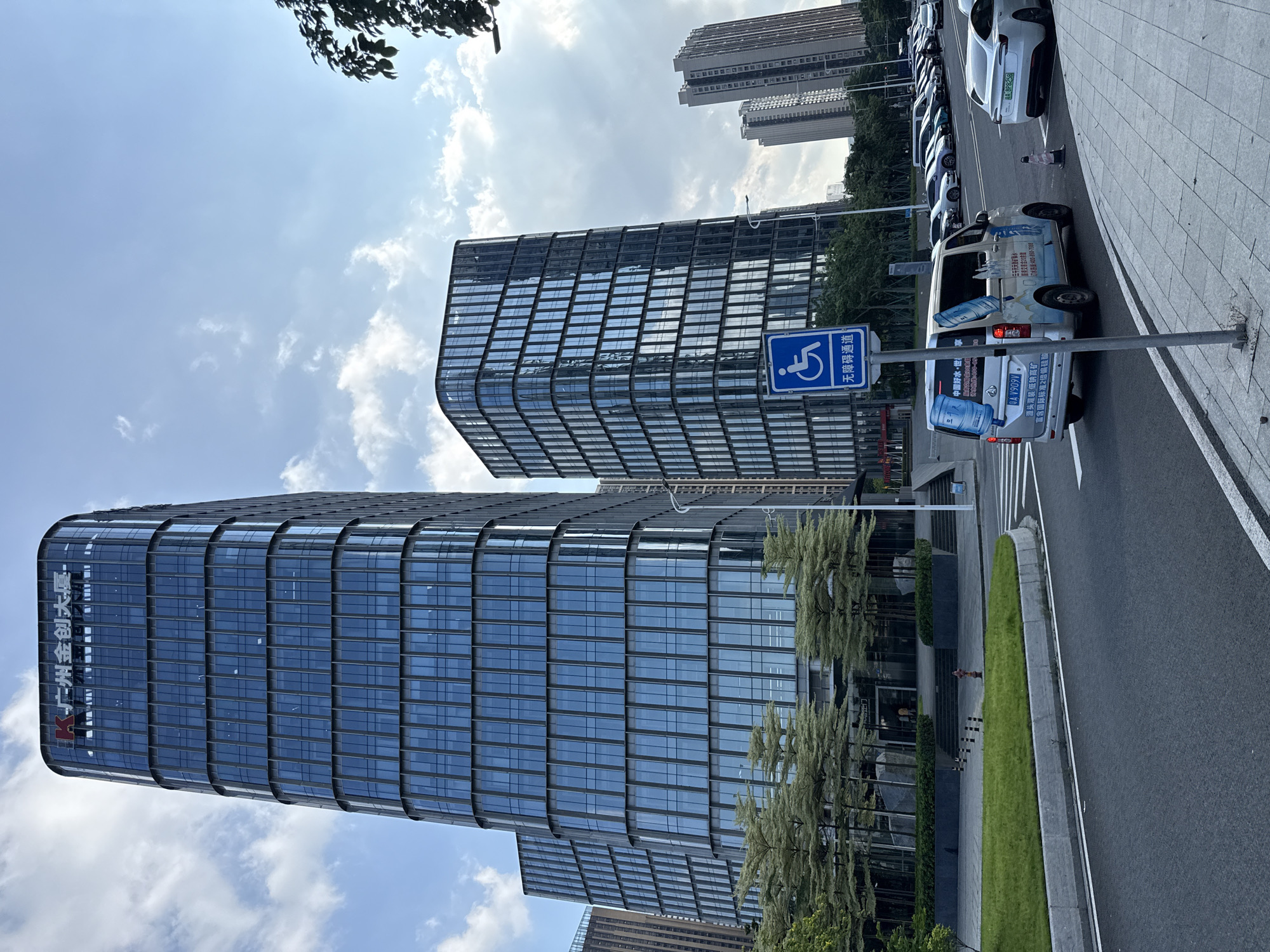 This is a vertical, eye-level, medium shot of a modern urban street scene in Haizhu District, China. The prominent subjects are two sleek, glass-facade office buildings, their reflective surfaces mirroring the sky. In the foreground, a row of parked cars, including a white electric vehicle and a white van adorned with water bottle graphics and Chinese text, lines the asphalt road. A blue sign with a white wheelchair symbol and Chinese characters indicating "Barrier-Free Access" stands near the van. The weather appears to be sunny with scattered clouds, suggesting it's daytime. Green landscaping, including grass and trees with feathery foliage, softens the hard edges of the buildings and road. The overall impression is one of a well-developed, modern business district. The visible text includes signage on the buildings, the van, and a parking permit on one of the cars. The text on the buildings reads "Guangzhou Jinchuang Building". The van has branding related to "China Good Water" and a helpline number. The electric car's license plate is visible.