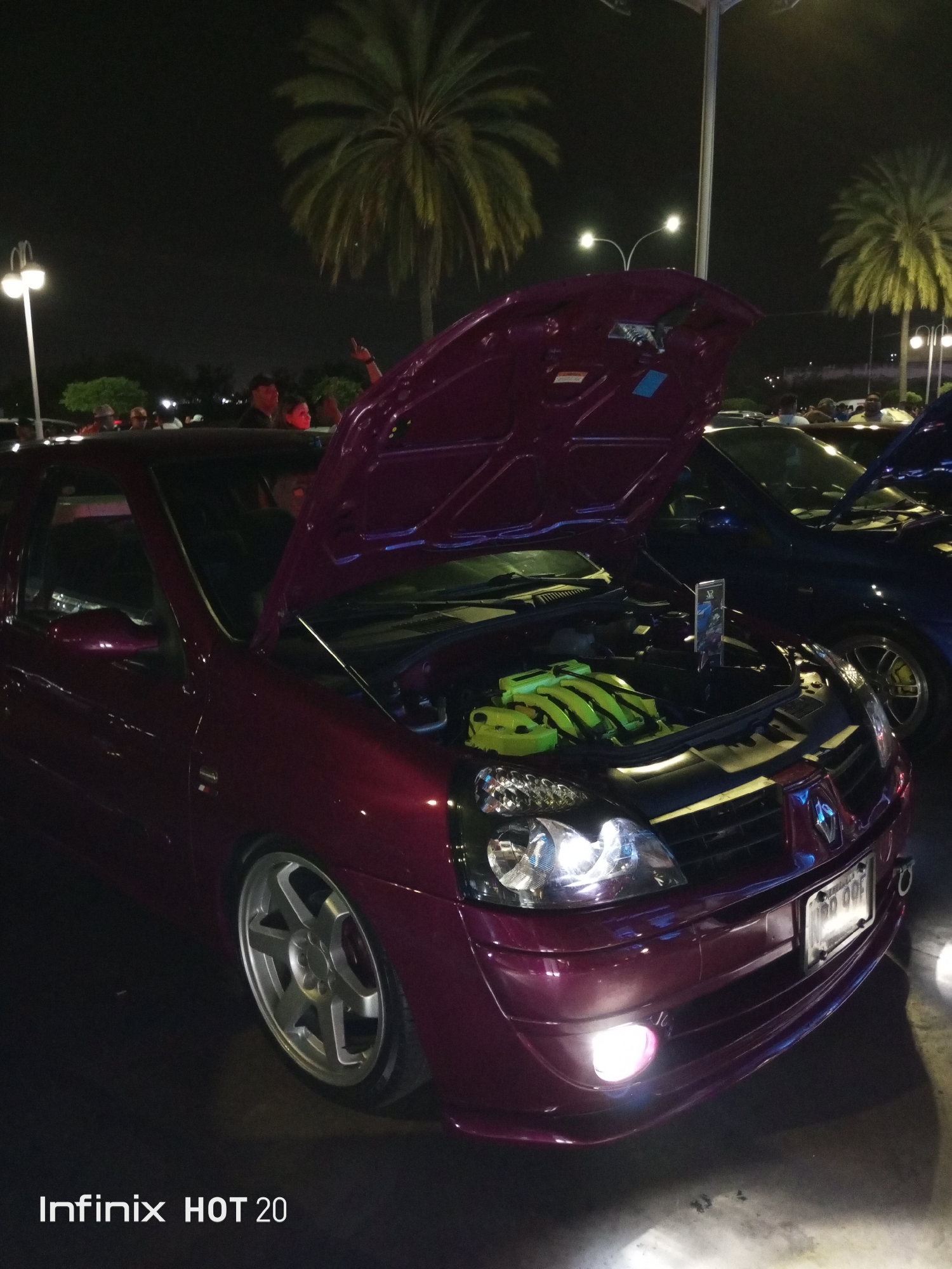 At night, a purple Renault Clio stands with its hood open, revealing a neon green engine manifold. Parked nearby is a blue vehicle with its hood also open. The Clio has aftermarket rims and an illuminated license plate. A street lamp is visible in the background, along with palm trees and people congregating around the cars. The "Infinix HOT 20" watermark indicates the image was taken with that device. The location is Porlamar, Venezuela.