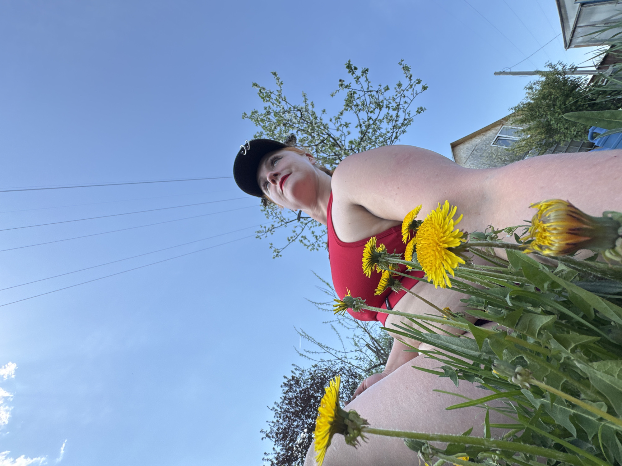 A low-angle shot shows a woman in a red top and a black cap with metal ring embellishments, looking upwards and to the left. She is surrounded by a cluster of dandelions in full bloom, with some plants closer to the camera and blurring the foreground.
In the background, a pale blue sky is visible, crossed by telephone or power lines. Sparse trees with light green foliage are seen, and behind the woman, a building can be partially seen, possibly a residence or small commercial structure. The location could be the city of Chaikovsky, Russia.
The woman's expression appears contemplative. She has fair skin and her red lipstick is noticeable. The lighting suggests it is daytime, likely in the early afternoon.