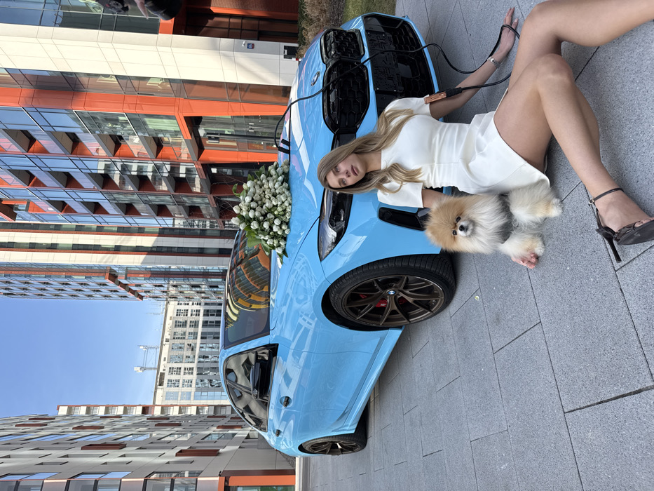 An outdoor scene in the Browary Warszawskie district of Warsaw, Poland, depicts a light blue BMW M3 G80 series sedan, a female individual, and a Pomeranian dog on a grey paved surface. The vehicle is parked with a large bouquet of white tulips positioned centrally on its hood. A red and black jumper cable is connected to the front grille area of the car and extends towards the female. The female, with long blonde hair, is seated on the paving stones, leaning on her left arm with her legs partially extended. She is wearing a white short-sleeved dress and dark high-heeled sandals. A red and black jumper cable clamp is attached to the cuff of her right sleeve, with the cable trailing towards her right hand. To her immediate left, a fluffy light brown Pomeranian dog wearing a grey harness is seated, looking directly forward. The background consists of modern high-rise buildings featuring facades with prominent orange-red panels and lighter beige sections, under a clear blue sky.