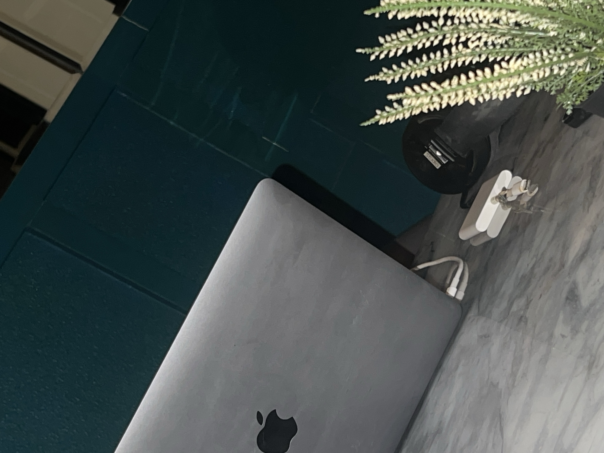 The rotated image presents an indoor scene featuring a silver laptop, a faux plant, and a white power adapter. The laptop, displaying the characteristic Apple logo on its lid, appears closed or partially closed, resting on a light-colored surface with a marble or granite pattern. A white power adapter is situated next to the laptop, with its cable connected, suggesting the laptop might be charging or ready for use.
Adjacent to these items is a prominent dark teal surface, possibly a piece of furniture or a wall panel, exhibiting subtle rectangular patterns or divisions. In the upper right portion of the image, a decorative faux plant with clusters of small, white, spike-like flowers emerges from a black pot. Faint white text, possibly reading "STOCk", is visible on the side of the pot.
The lighting in the image is dim, indicating an indoor environment, likely during evening hours or in a moderately lit room. In the background, to the far left, fragments of white and dark horizontal slats are discernible, possibly part of window blinds or another furniture item. The arrangement suggests a stationary setup on a desk or table, potentially within a home or office in Abuja, Nigeria. No people are present, and the scene captures a still life of technological and decorative items.