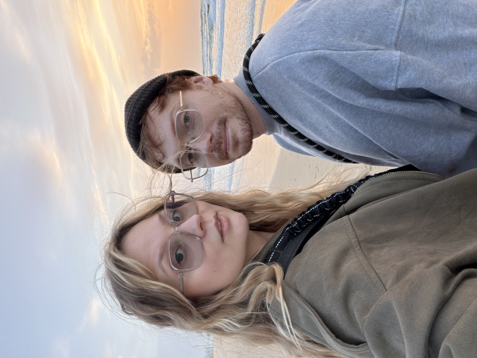 A young adult female and male are featured in a waist-up, direct-to-camera photograph on a sandy beach in Tel-Aviv, Israel, during sunset. The sky displays hues of orange, yellow, and light blue with cloud formations. The Mediterranean Sea with gentle waves is visible in the background. The female, on the left, has long wavy blonde hair and wears large, square-framed, light-tinted glasses and an olive-green zippered jacket. A black and white speckled strap crosses her left shoulder. She looks at the camera with a slightly open mouth. The male, on the right, has reddish-brown hair and a light beard, wearing a black knit beanie, rectangular gold-framed clear glasses, and a grey crew-neck sweatshirt. A black rope-like strap with white flecks crosses his right shoulder. He looks at the camera with a neutral expression.