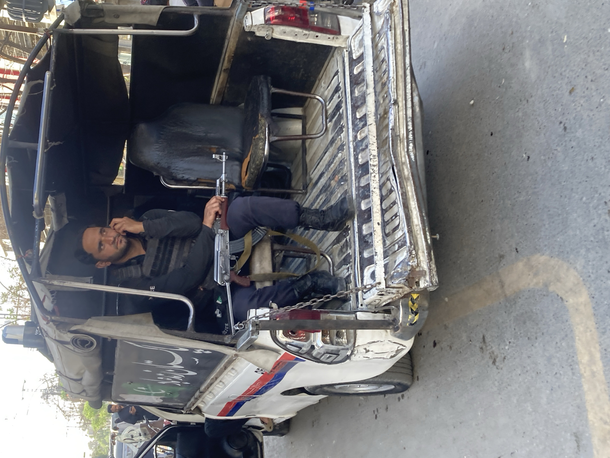 A man sits in the back of a white auto-rickshaw, holding a rifle in his hands. The rickshaw appears to be damaged, with visible dents and scratches on its body. The vehicle is decorated with white, blue, and red stripes, which could indicate it's used for official purposes. The man is wearing a black vest over his clothes and black boots, suggesting a uniform of some kind. The scene is set in what appears to be Quetta City Tehsil, Pakistan, as evidenced by the distinctive auto-rickshaw design common in the area. He is also speaking on the phone.