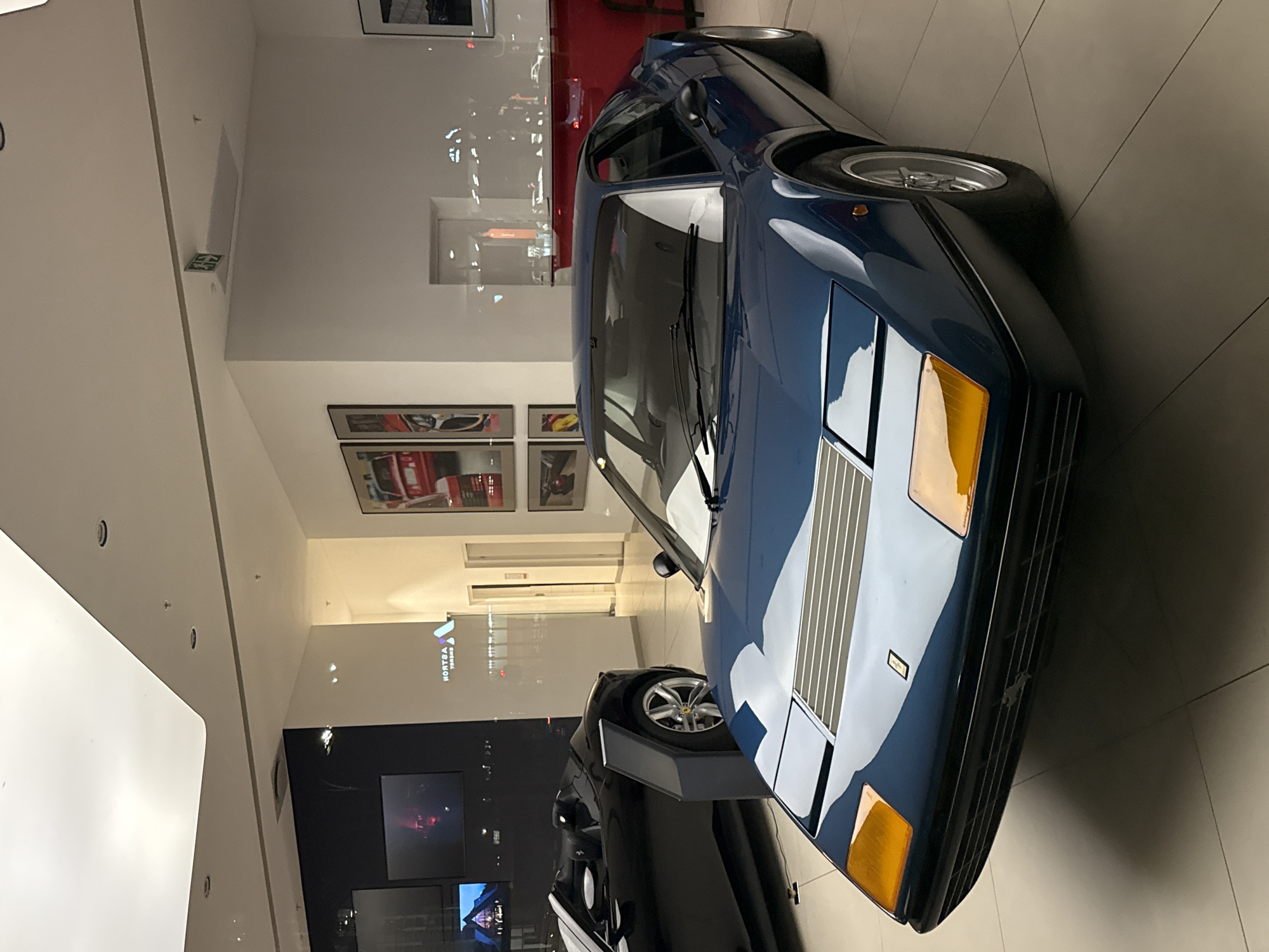 The image depicts the interior of a brightly lit automotive showroom or gallery, identified as being in South Africa, specifically within the city of Cape Town, based on the reflected "ASTRON ENERGY" logo visible on a wall.
In the foreground, a dark blue vintage Ferrari Berlinetta Boxer (likely a 512 BB model) is positioned centrally, facing slightly left. The vehicle exhibits a flat front design with orange rectangular covers where retractable headlights are located, a front bonnet featuring horizontal grille slats, and a small Ferrari emblem. It rests on a light-colored tiled floor and is stationary. To its left, partially obscured in the mid-ground, is a black, open-top Ferrari convertible, resembling a Monza SP1 or SP2, with a visible silver multi-spoke wheel and distinct body lines; it is also stationary.
The environment consists of predominantly white walls, with a section on the far left being dark (black) and featuring screens displaying illuminated content. Framed automotive artwork, primarily depicting red sports cars, hangs on the white wall to the right. A mirrored "ASTRON ENERGY" logo is reflected on the white wall in the center-left background. An illuminated green "EXIT" sign with a left-pointing arrow is visible on the upper right wall-ceiling junction. A red counter or display unit is partially visible in the far right background. The ceiling is white with recessed lighting and a curved architectural feature on the left. Reflections of what appear to be distant urban lights are visible on glass surfaces in the background. No people are present or visible within the frame, and there are no ongoing actions or interactions.