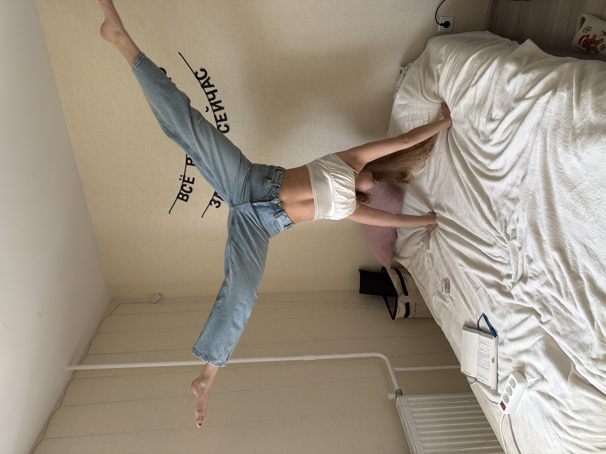 A young woman in a white top and blue jeans is performing a gymnastic split with her legs extended upwards towards the ceiling of a room. Her hands are touching the edge of a bed covered with a white blanket. The room has light-colored walls with Russian text on one wall. The bed has a pink pillow and a storage bag next to it. A notebook and a power strip are on the bed. A radiator is visible near the bed. The location is likely Izhevsk, Russia, given the text on the wall.