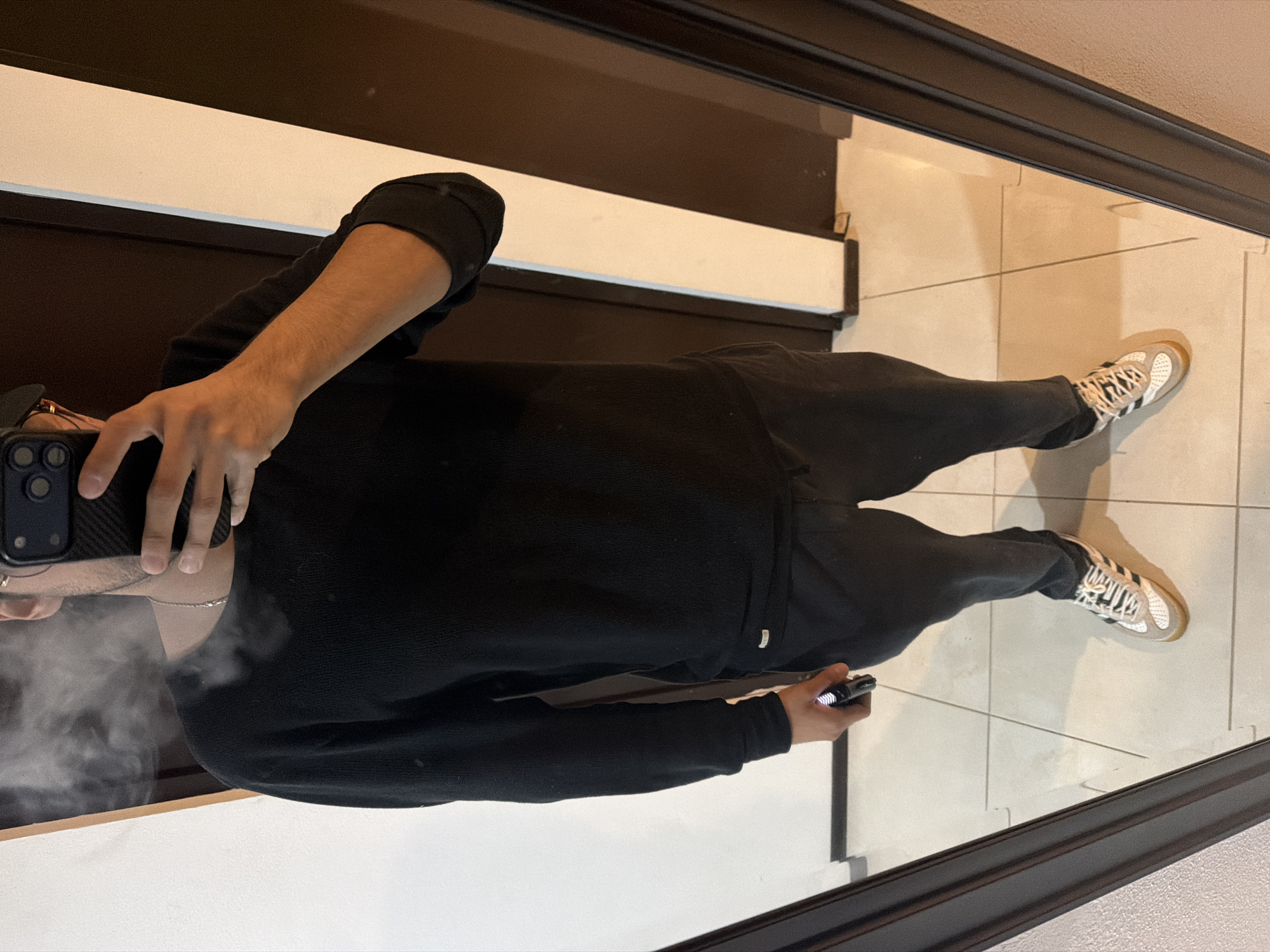 A man takes a mirror selfie in a bathroom with tile flooring and a black-framed mirror. The man wears black sweatpants, a black sweater, and white Adidas Samba sneakers. He holds a black cell phone in his right hand, and another in his left. A haze of fog is present around his lower face. The location is San Andrés Cholula, Mexico.